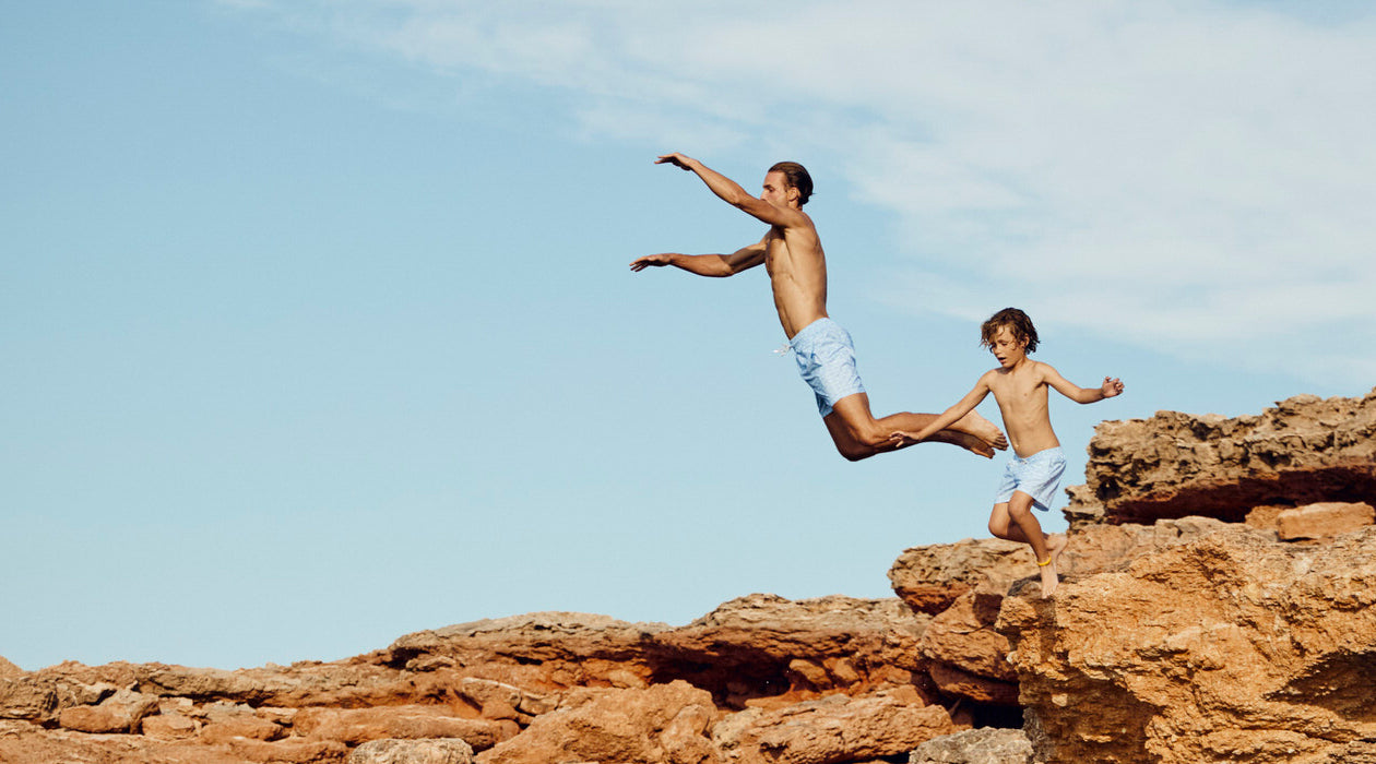 Father and son jump off rocks into water wearing designer swim shorts