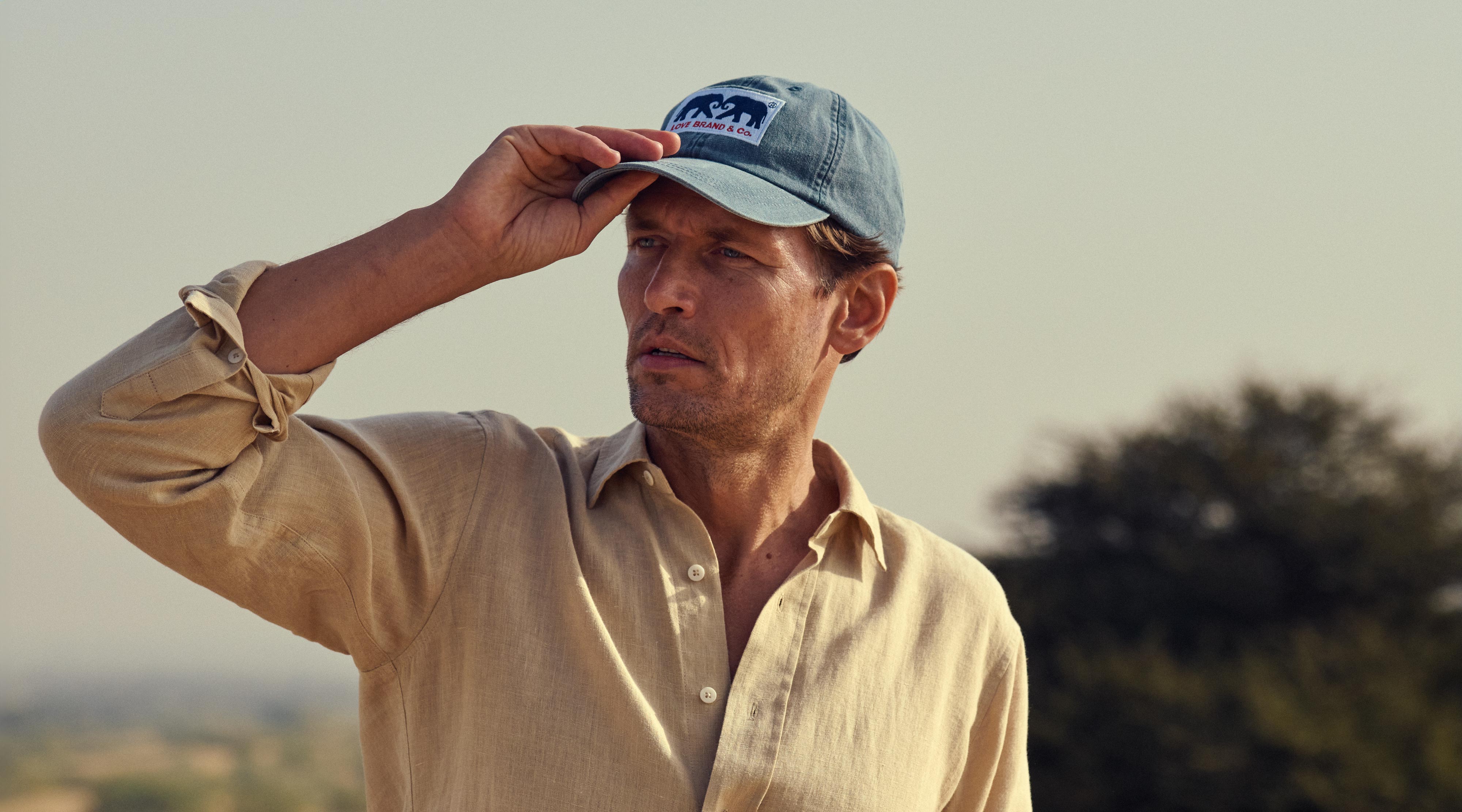 Model wears navy baseball cap and brown linen shirt