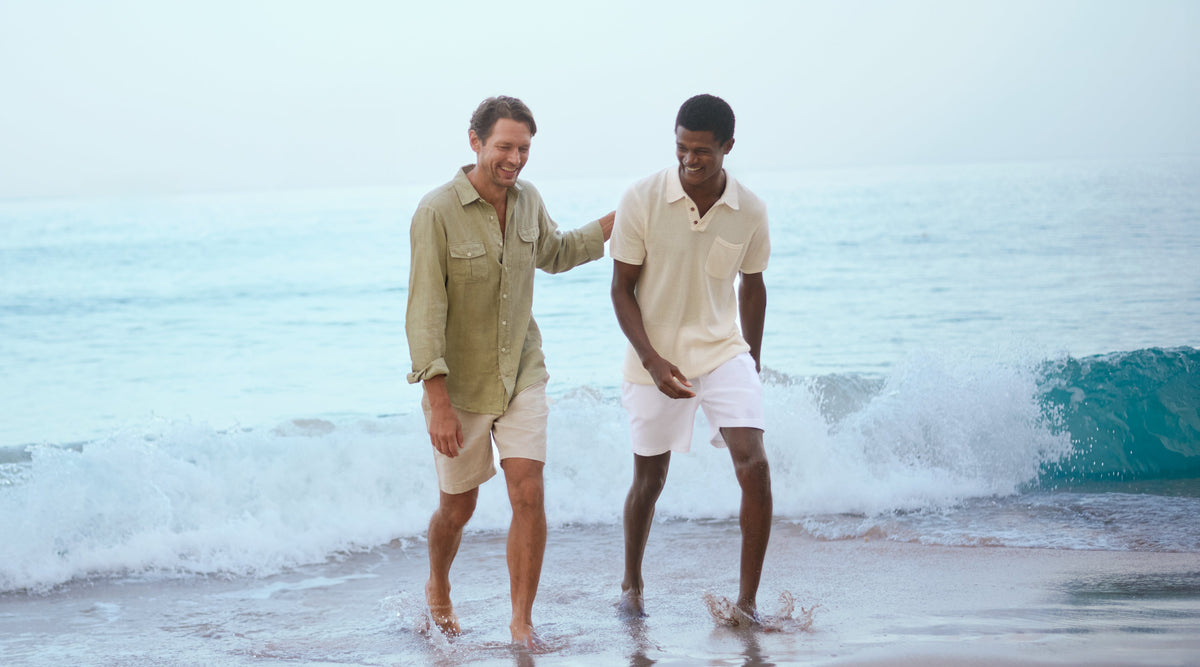 Man wears a green linen shirt and beach shorts next to another man wearing a knitted cotton polo shirt walking out of the ocean water