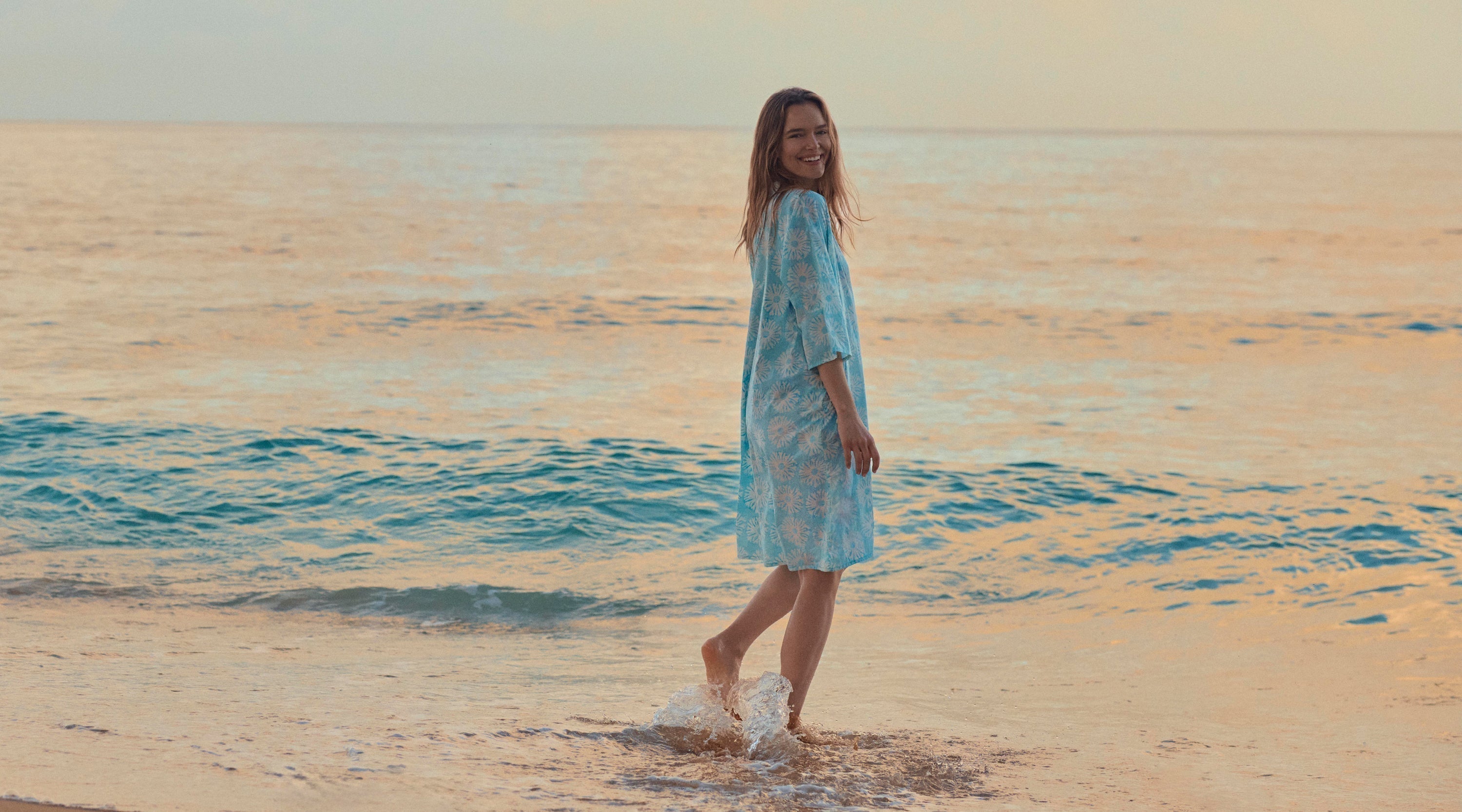 Woman wears blue beach dress while walking along the shore at sunset
