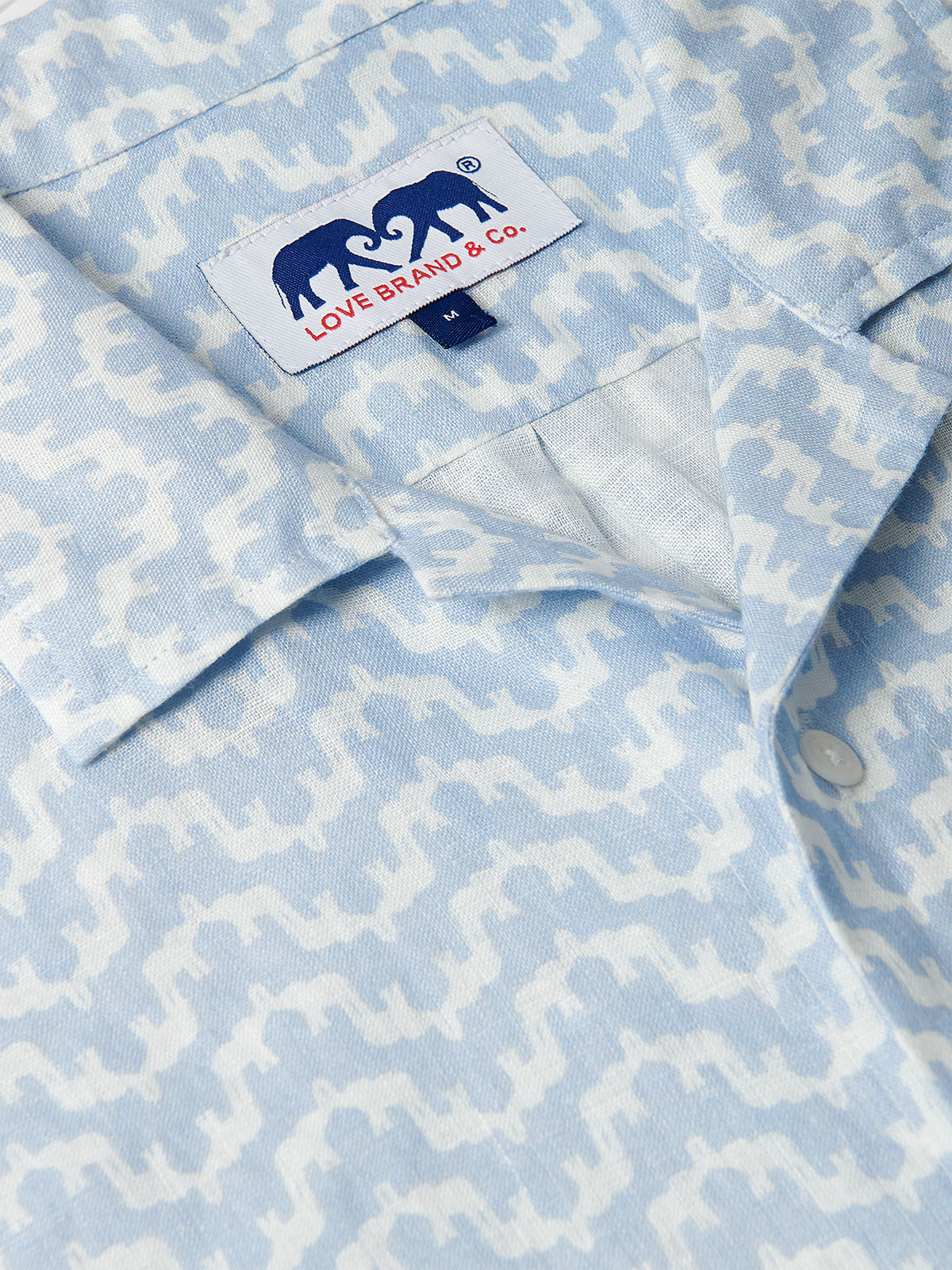 Close-up of Men's Elephant Palace Sky Arawak Linen Shirt. Men's linen shirt featuring light blue and white elephant and palace print, button details, and "Love Brand & Co." label with two facing elephants.