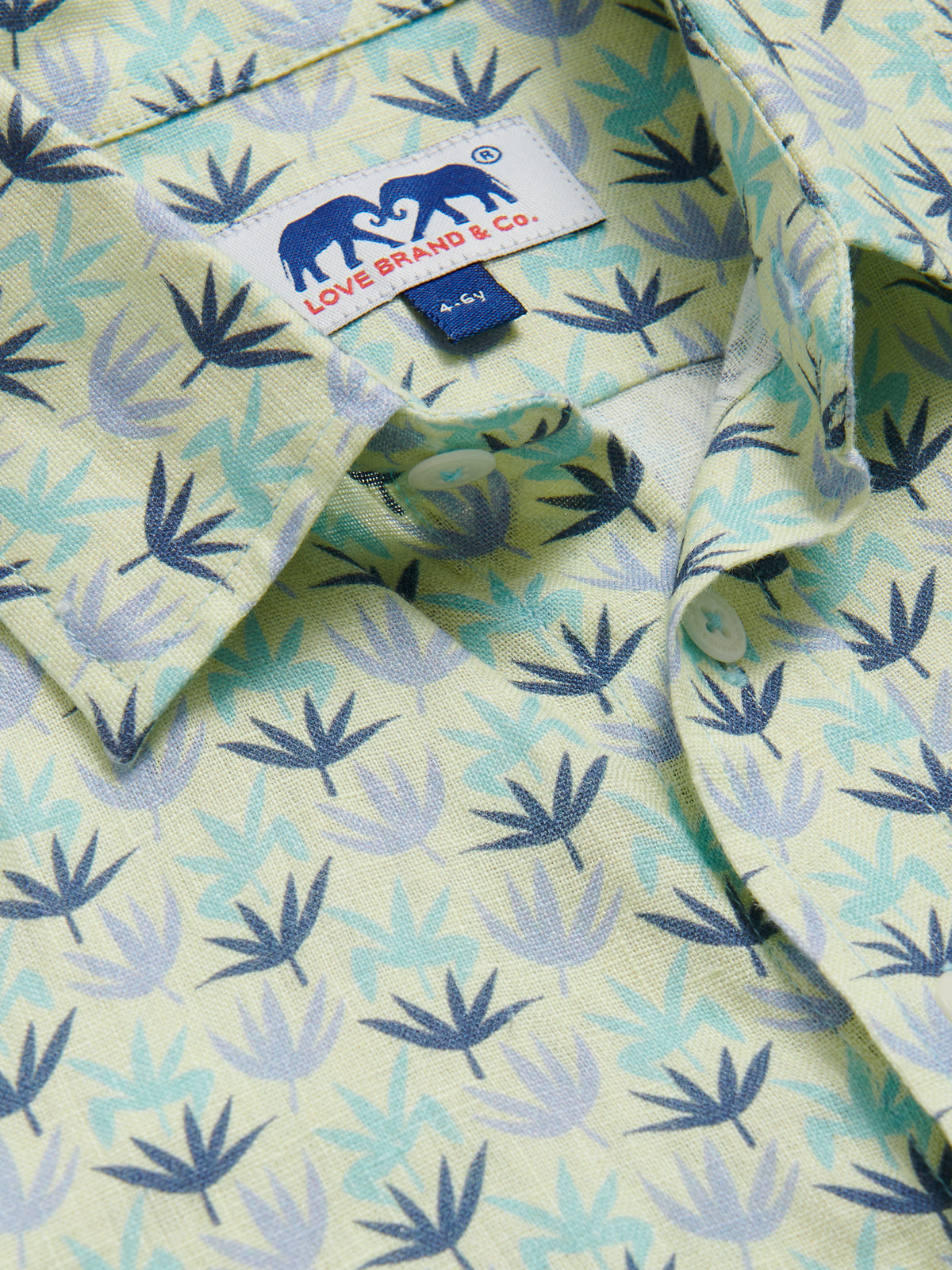 Close-up of the Boys Palm Sugar Abaco Linen Shirt featuring a tropical fan palm design in sky blue, aqua green, and deep blue on a limoncello yellow base, with a visible Love Brand & Co. label.
