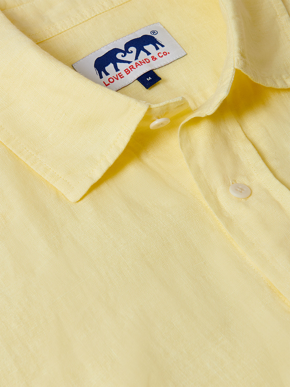 Close-up of Men's Limoncello Manjack Linen Shirt. Men's linen shirt showing a light yellow fabric with a collar, button details, and a tag from Love Brand & Co.