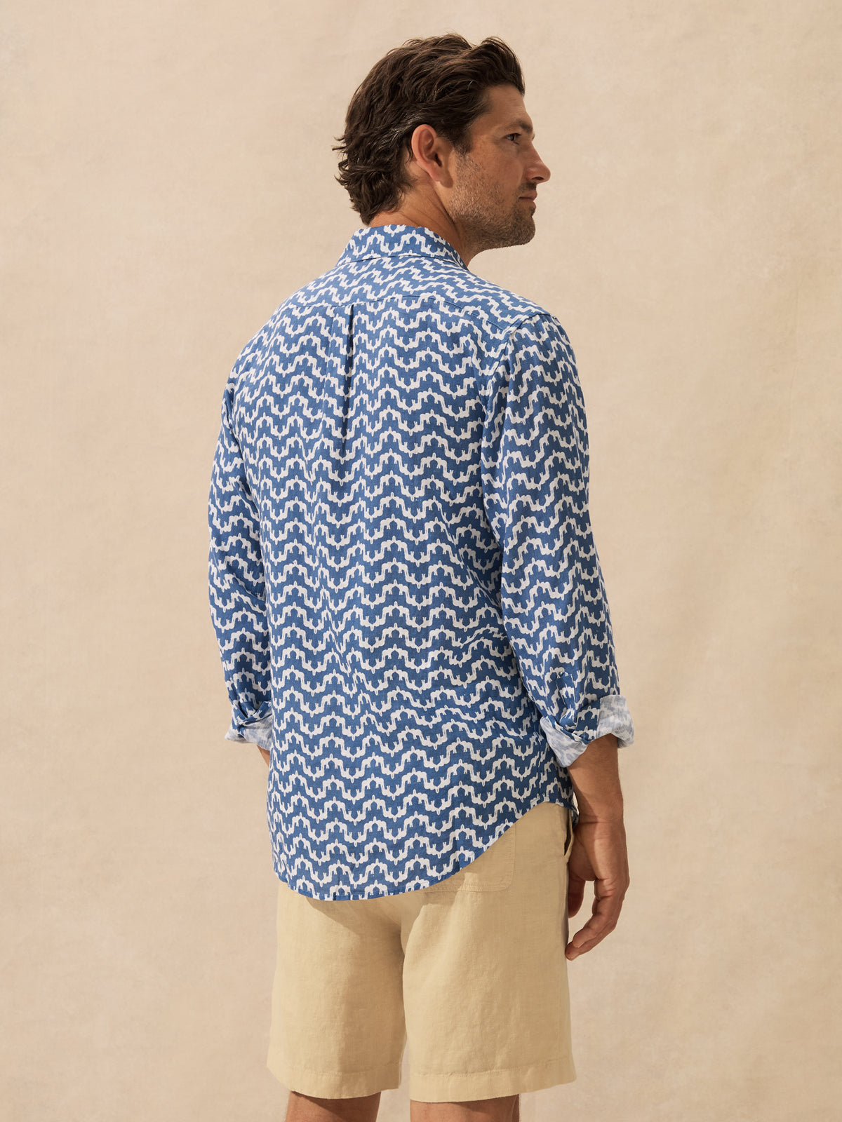 A man wearing a blue and white patterned long-sleeve button-down shirt and beige shorts, shown from a side-back angle against a neutral beige backdrop.