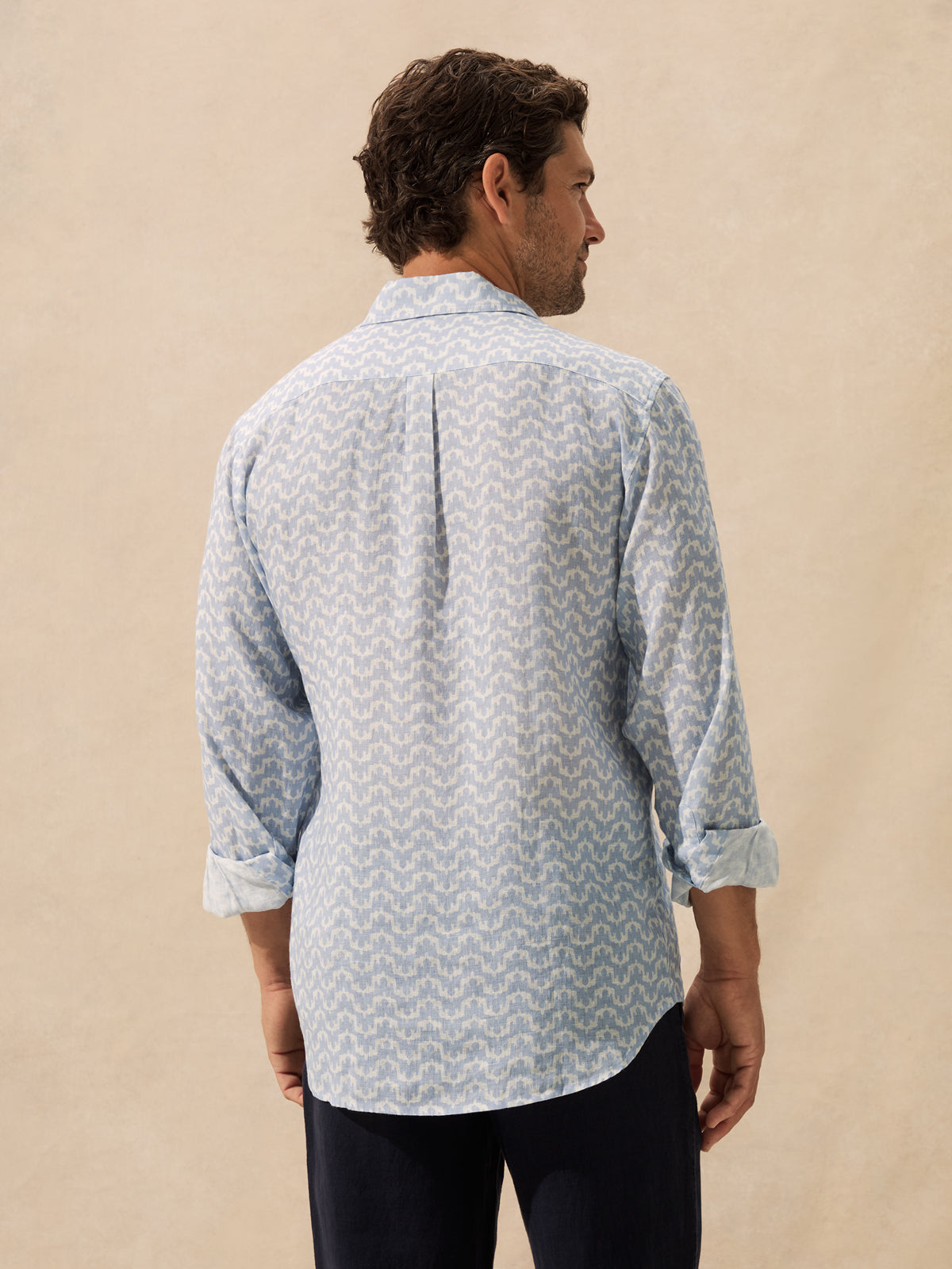 A man shown from the back wearing a light blue and white patterned button-down shirt standing against a textured beige background.