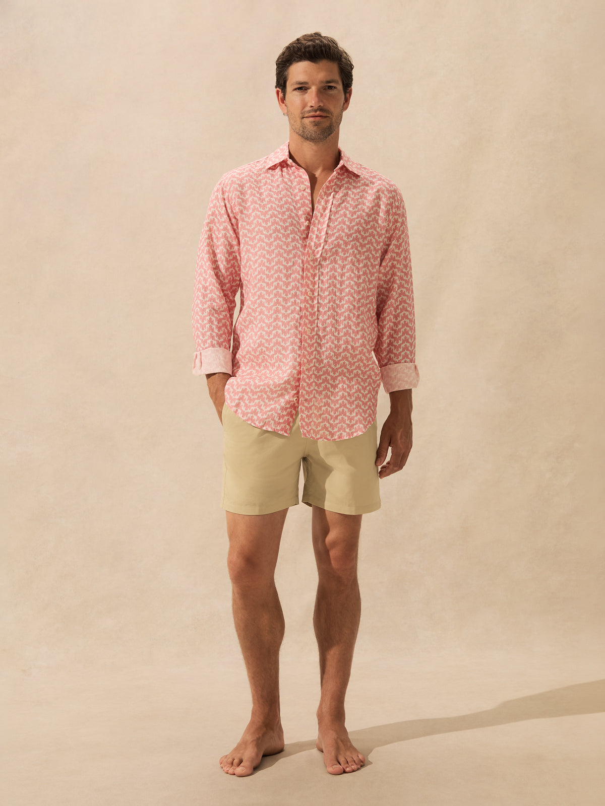 A man stands barefoot against a warm beige studio background, wearing a pink and white patterned long-sleeve button-up shirt and light beige swim shorts.