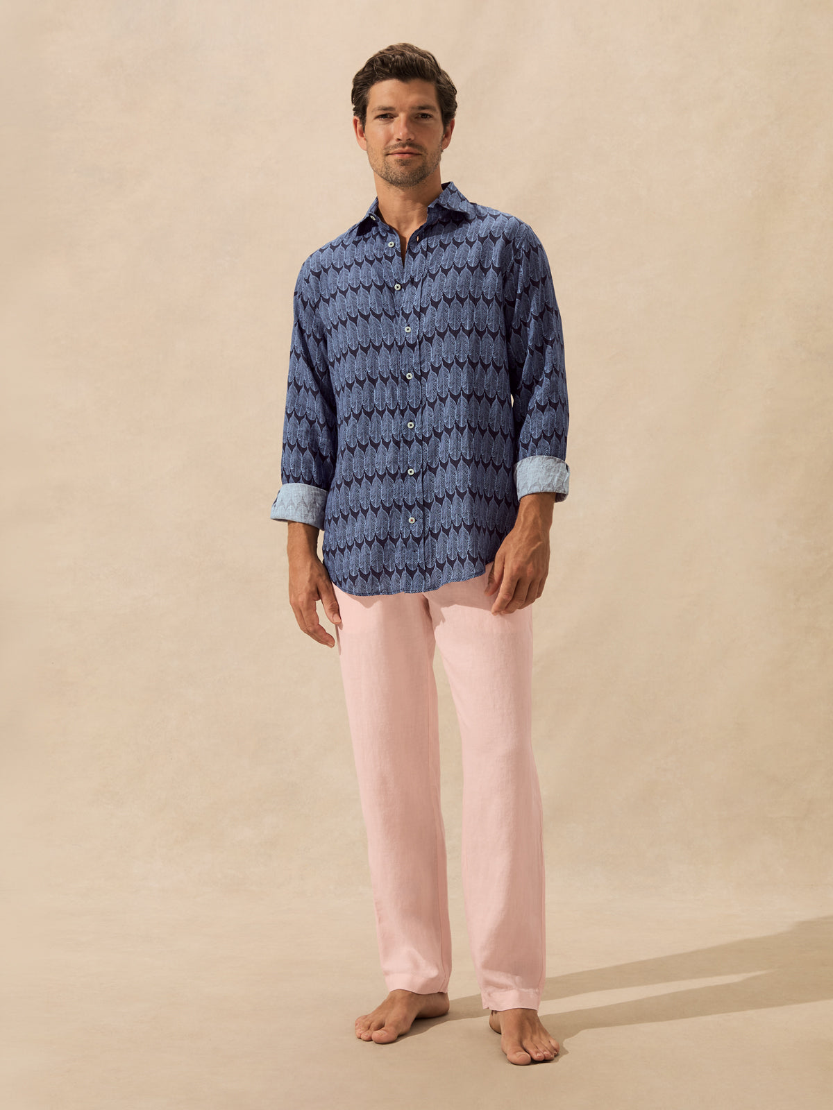 A man stands barefoot against a warm beige studio backdrop, wearing a blue patterned long-sleeve button-up shirt with light blue rolled cuffs and light pink pants.