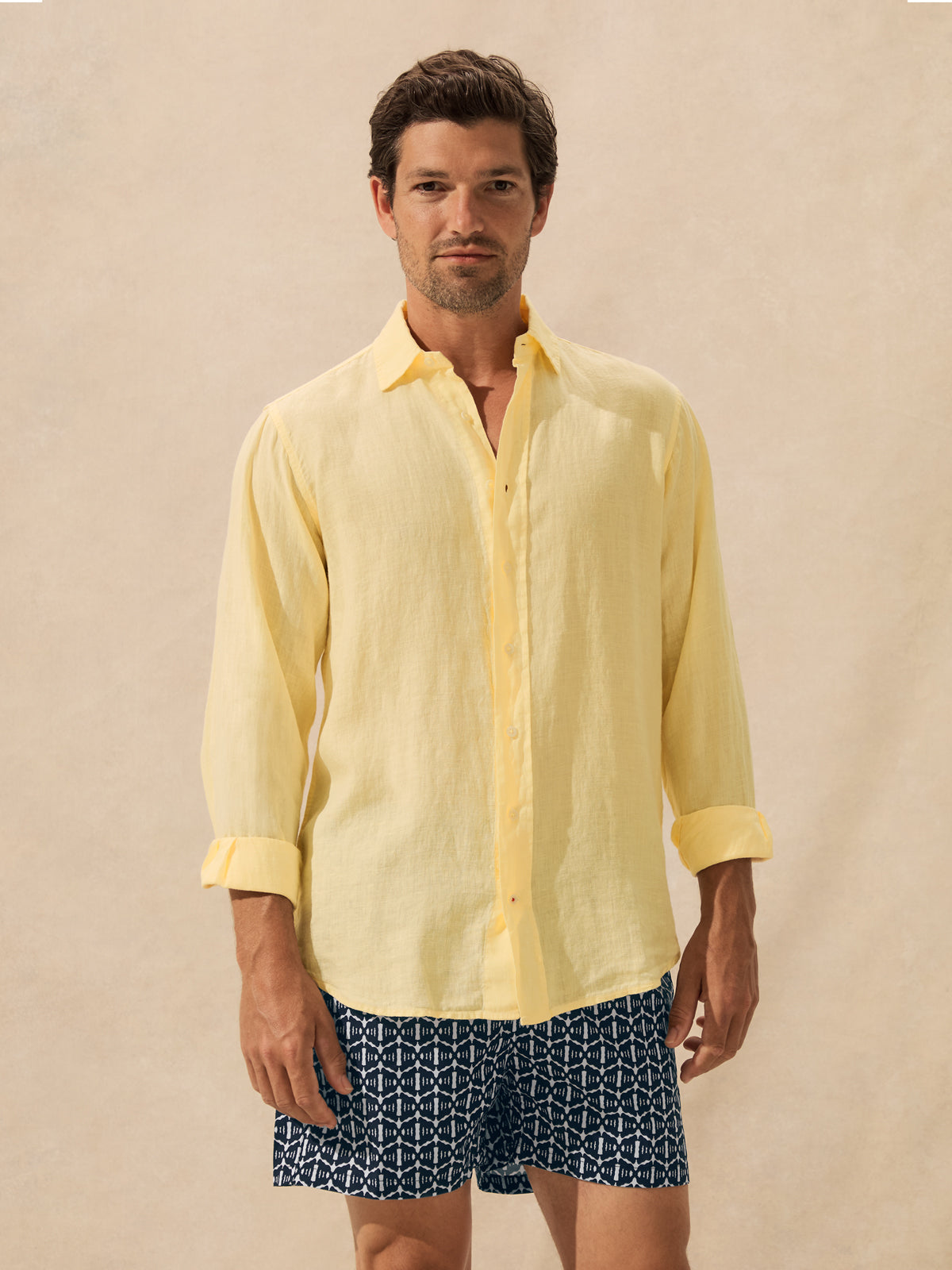 A man wearing a pale yellow long-sleeve linen button-up shirt and navy blue patterned shorts, standing against a warm beige studio background.