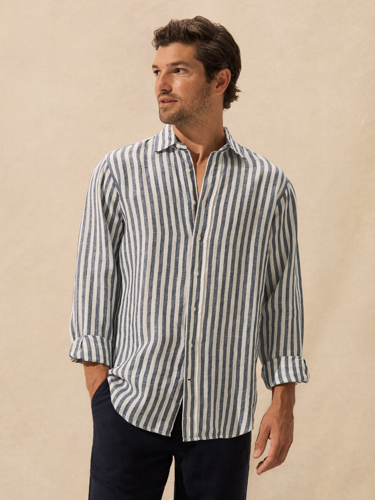 A man wearing a long-sleeve button-up shirt with vertical navy and beige stripes, paired with dark trousers, standing against a neutral beige background.