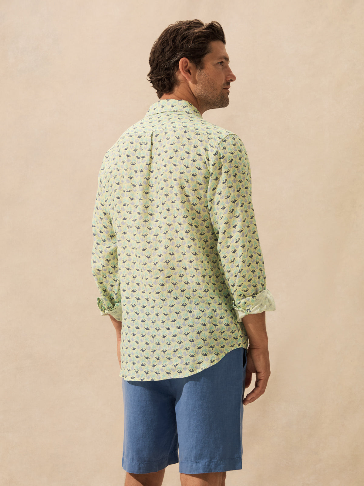 A man shown from the back wearing a light green long-sleeve button-up shirt with a small repeating leaf print and blue shorts, standing against a plain beige wall.