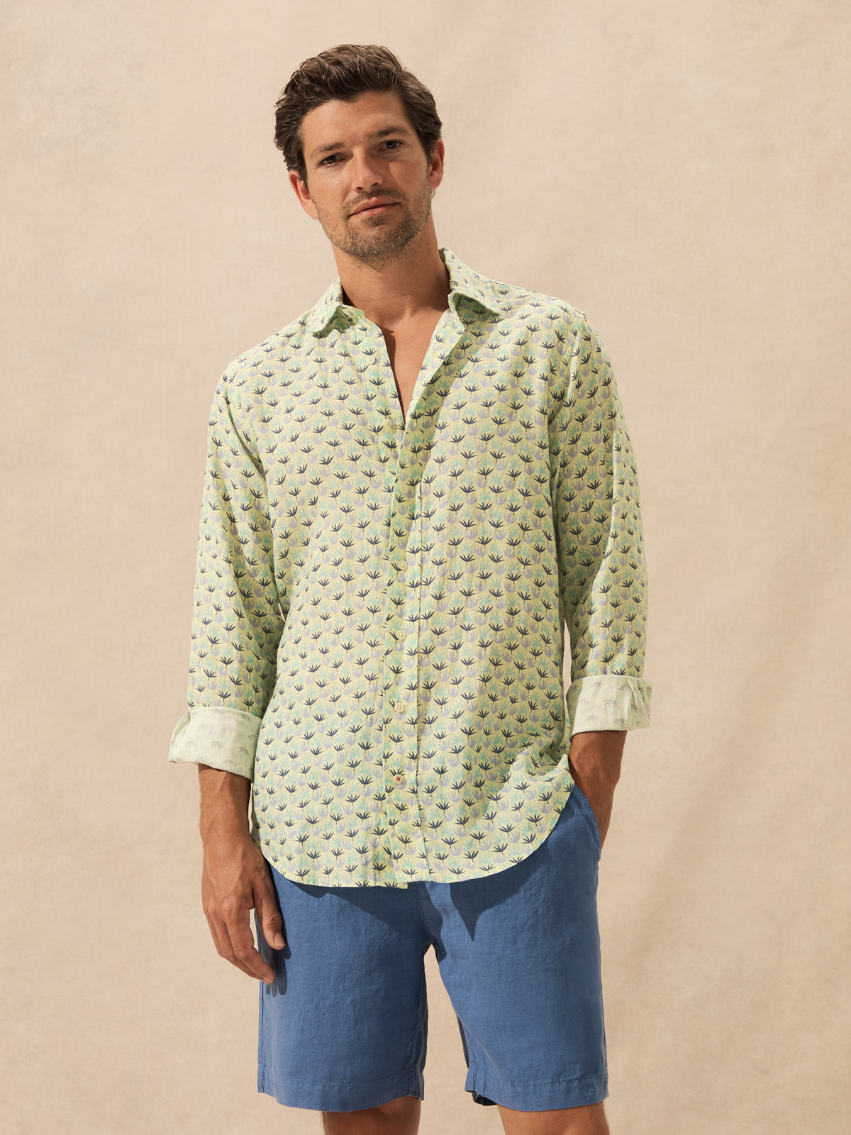 A man wearing a light green long-sleeve button-up shirt with a small repeating floral pattern and blue shorts, standing against a plain beige studio background.
