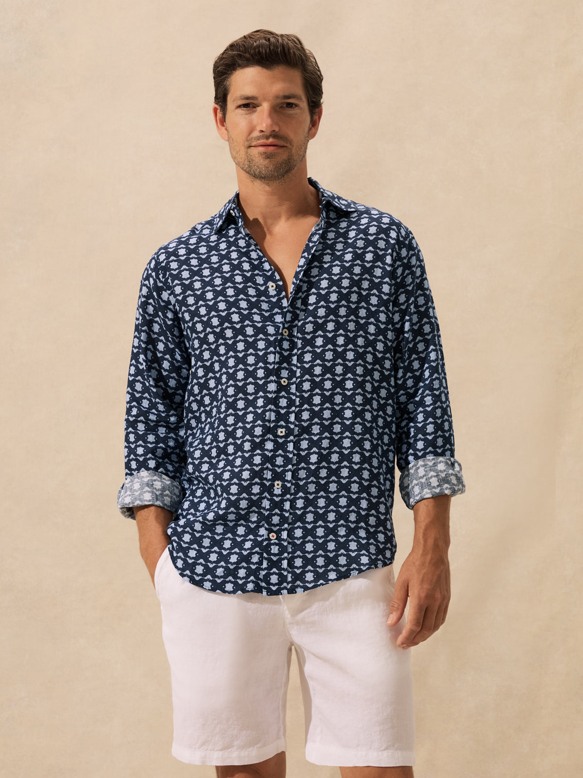 A man wearing a navy blue long-sleeve button-down shirt with a white geometric pattern, paired with white linen shorts, standing against a textured beige background.
