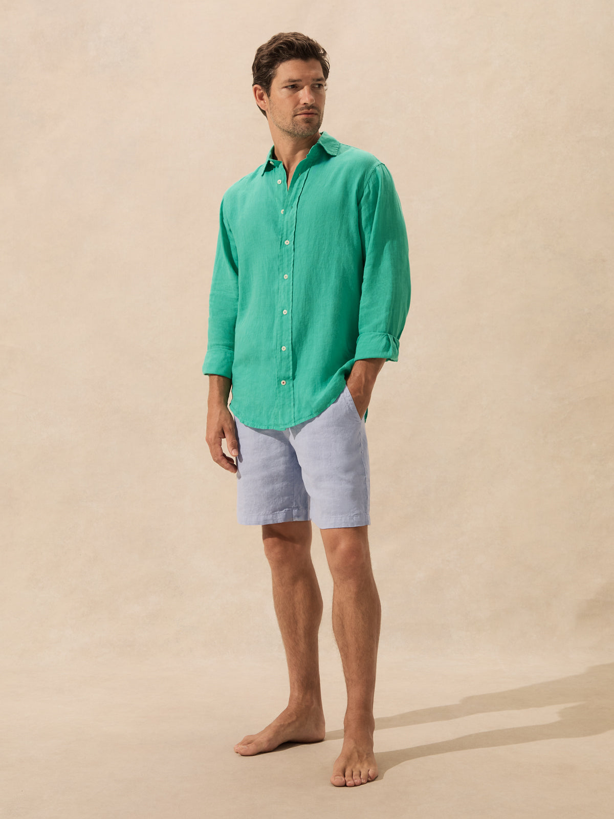 A man stands barefoot against a warm beige textured backdrop, wearing a bright green long-sleeve button-up shirt and light blue shorts.