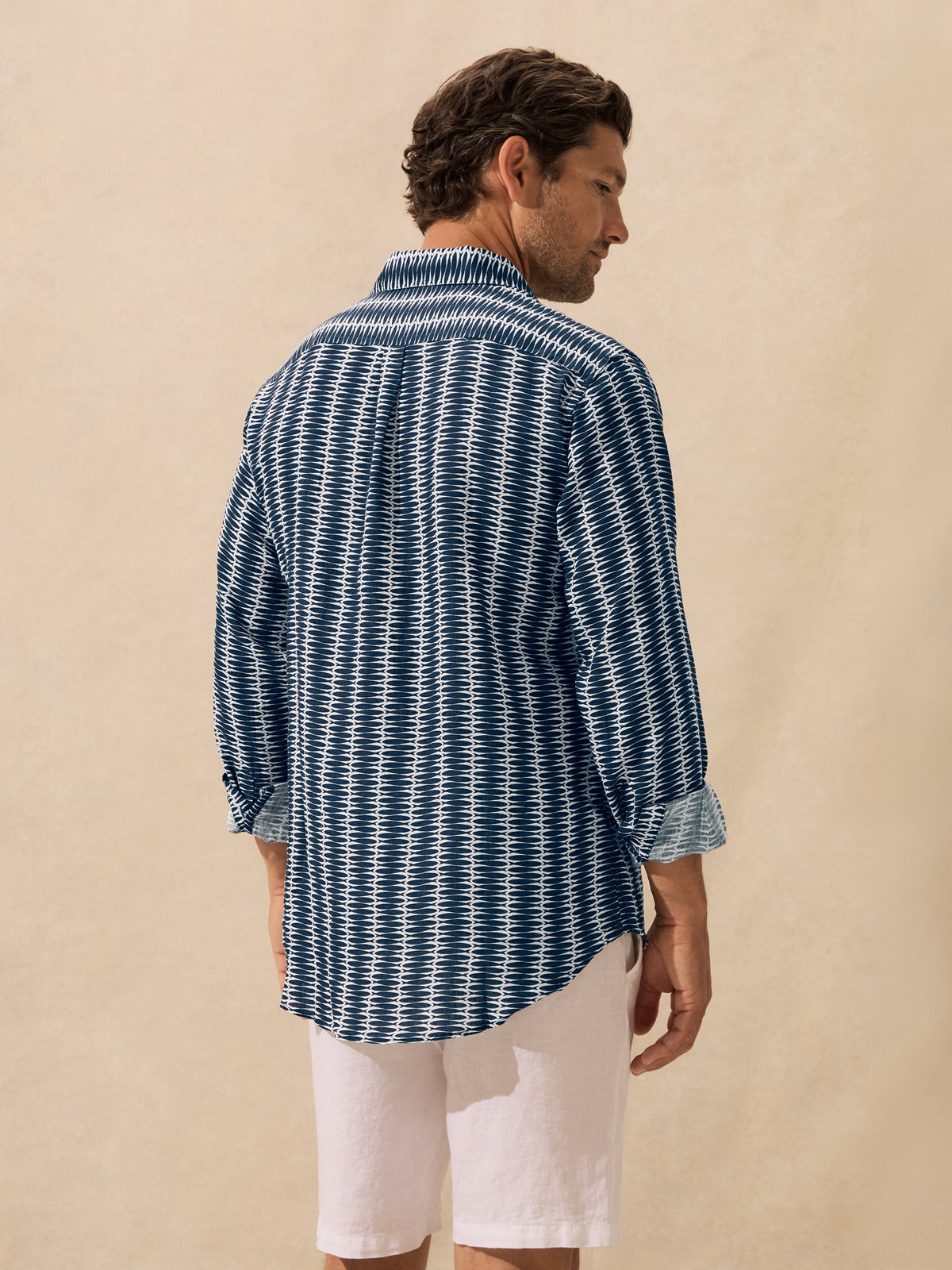 A man shown from the back wearing a blue and white patterned button-down shirt standing against a textured beige background.