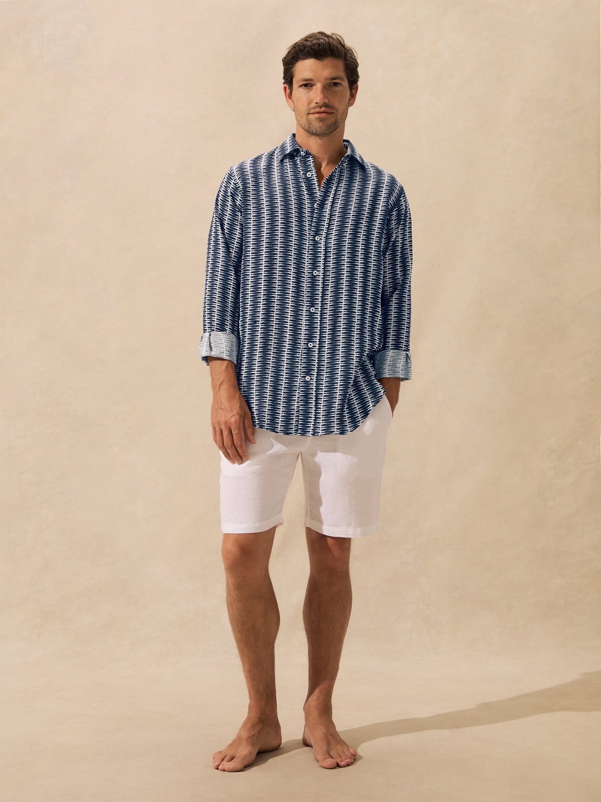 A man stands barefoot against a warm beige textured backdrop, wearing a long-sleeve button-up shirt with a blue and white geometric pattern and white shorts.