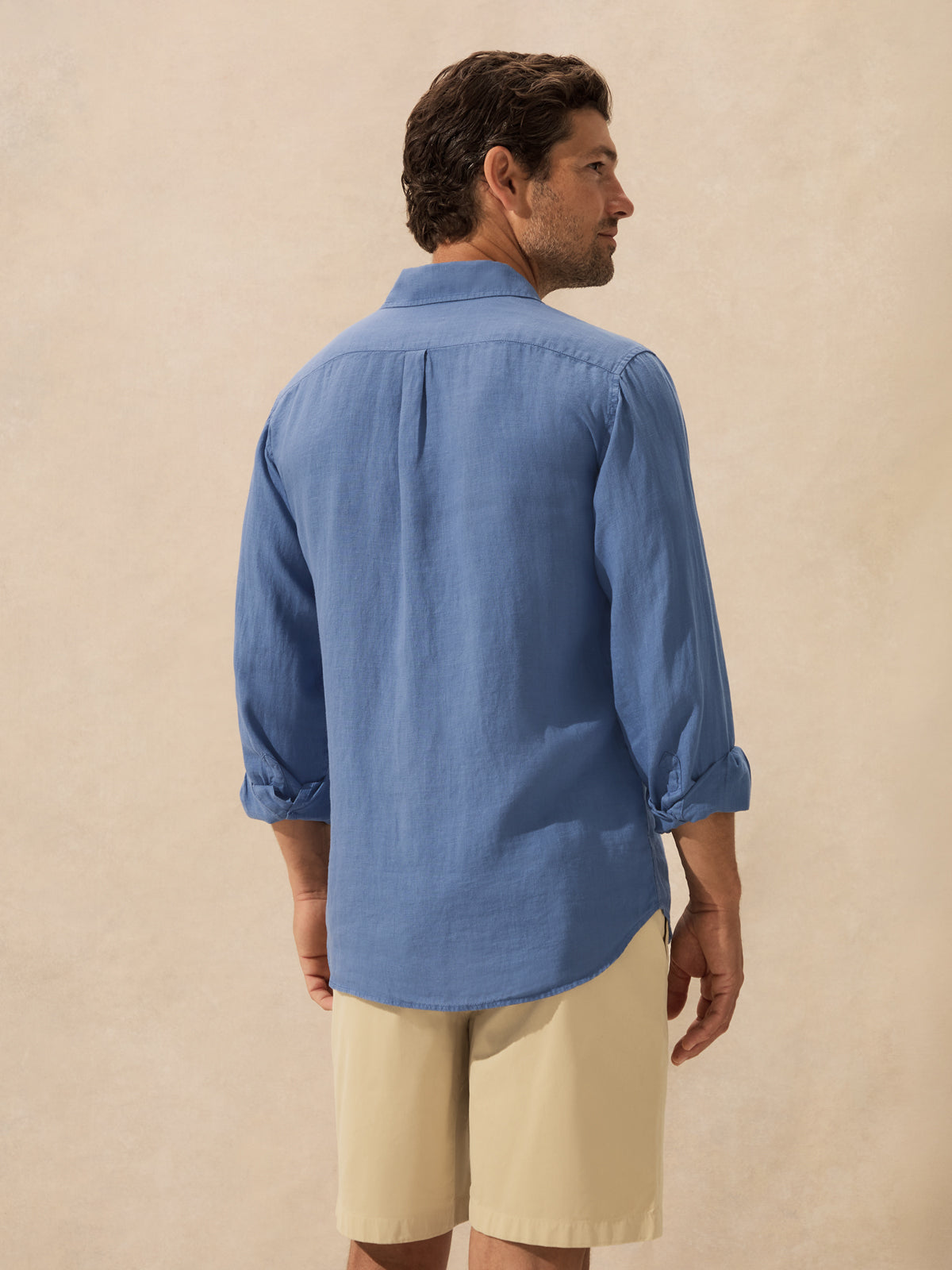 A man shown from the back wearing a light blue long-sleeve button-up shirt standing against a textured beige background.
