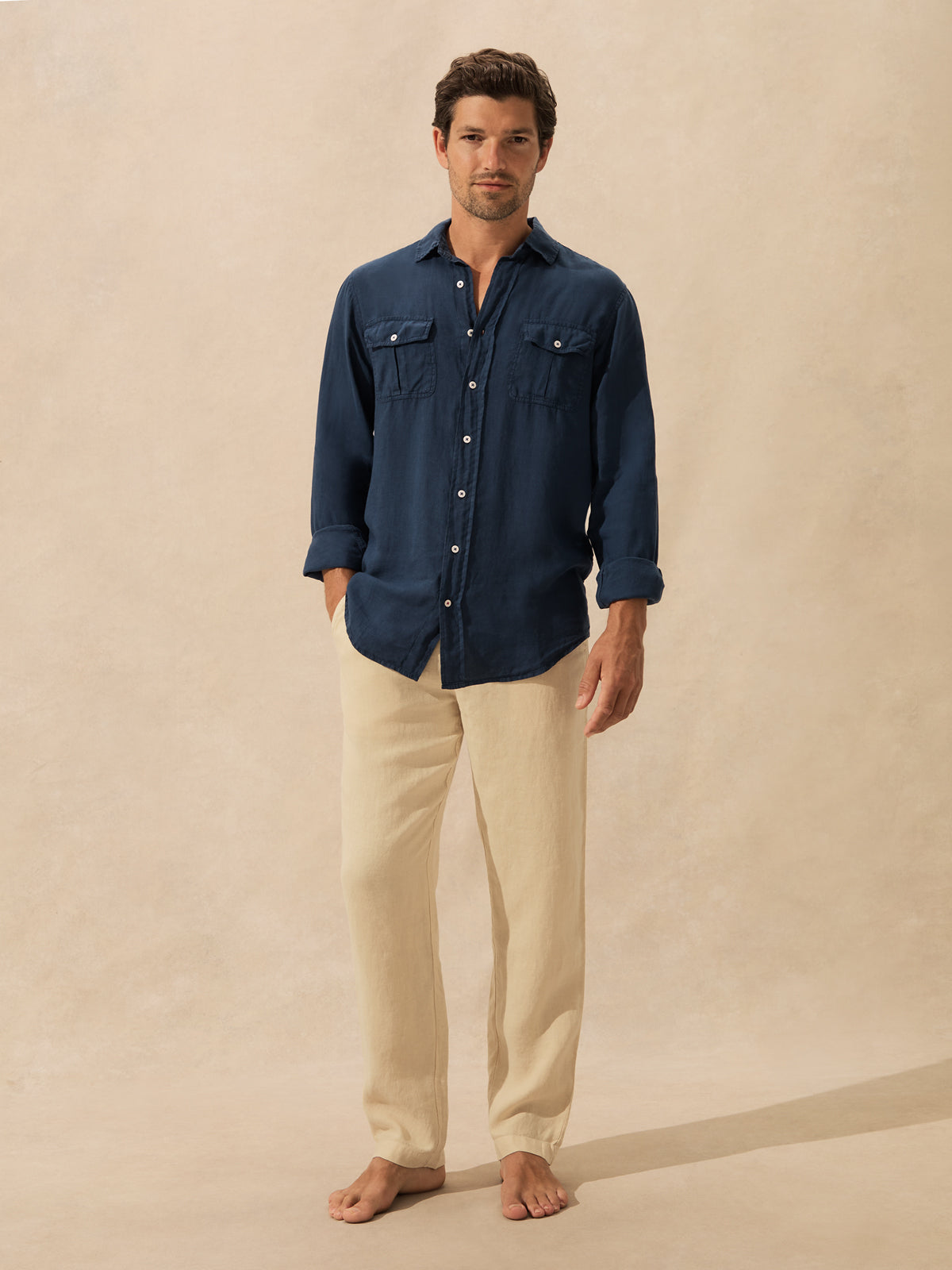 A man stands barefoot against a warm beige textured backdrop, wearing a dark navy long-sleeve button-up shirt with chest pockets and light beige pants.