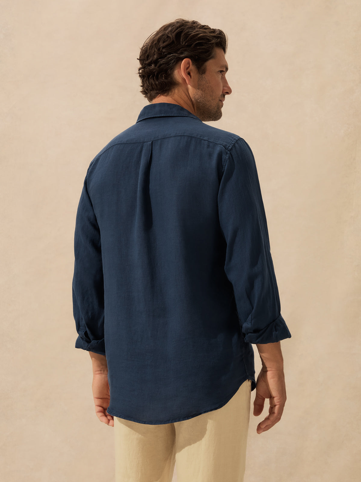 A man wearing a navy blue long-sleeve button-down shirt and beige trousers, shown from a rear three-quarter view against a textured beige background.
