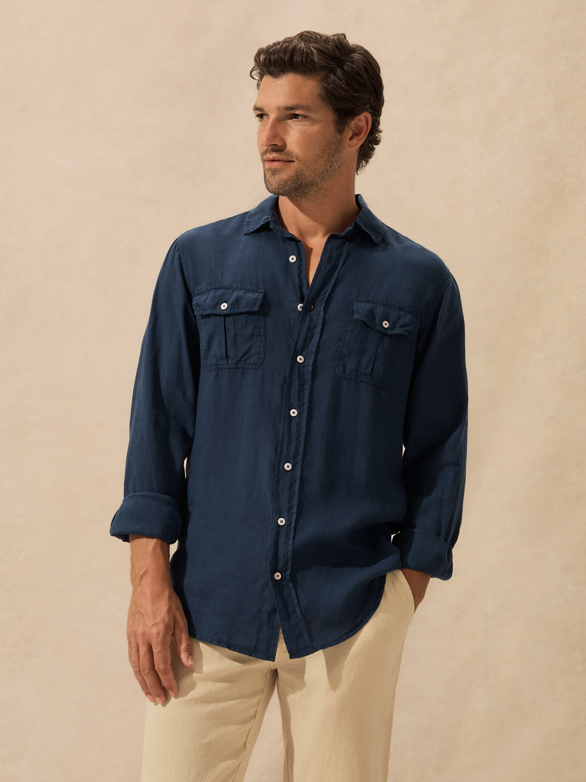 A man wearing a dark navy blue long-sleeve button-up shirt with two chest pockets and beige pants, standing against a textured beige background.