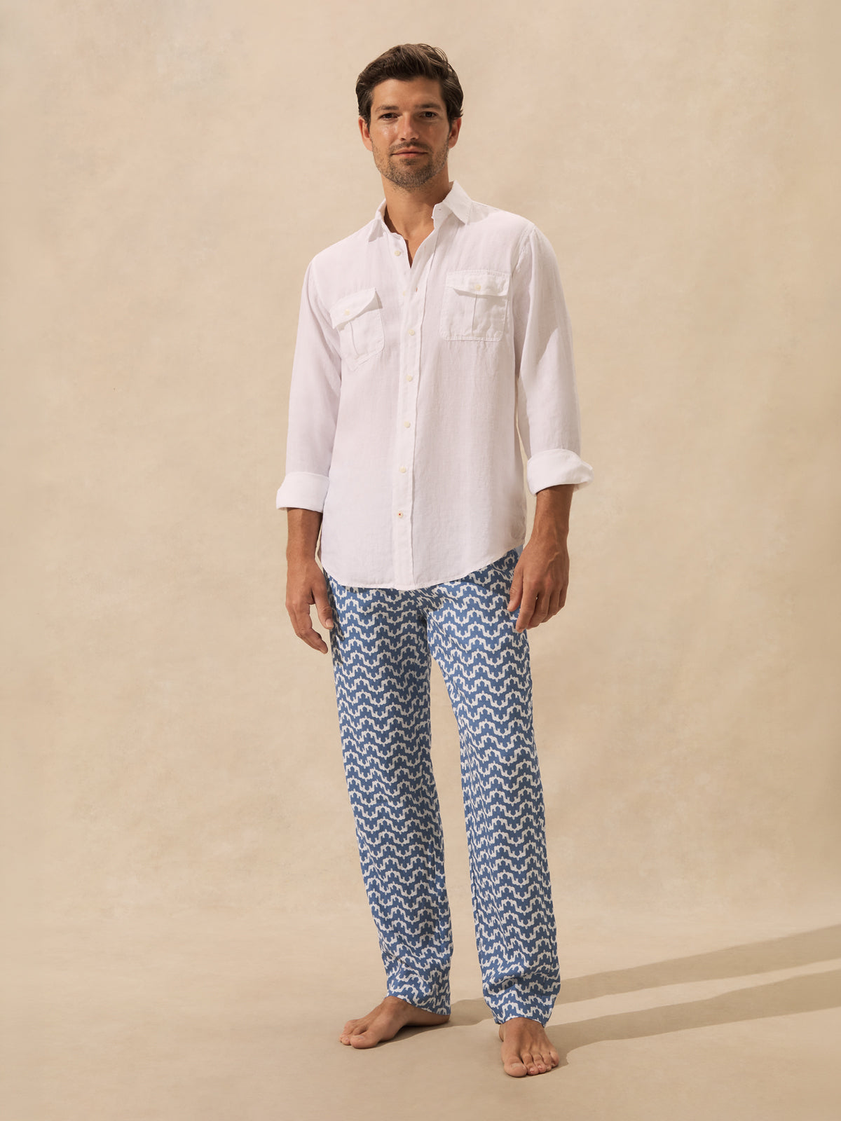 A man stands barefoot against a warm beige studio backdrop, wearing a white long-sleeve button-up shirt and blue patterned pants with a white geometric print.