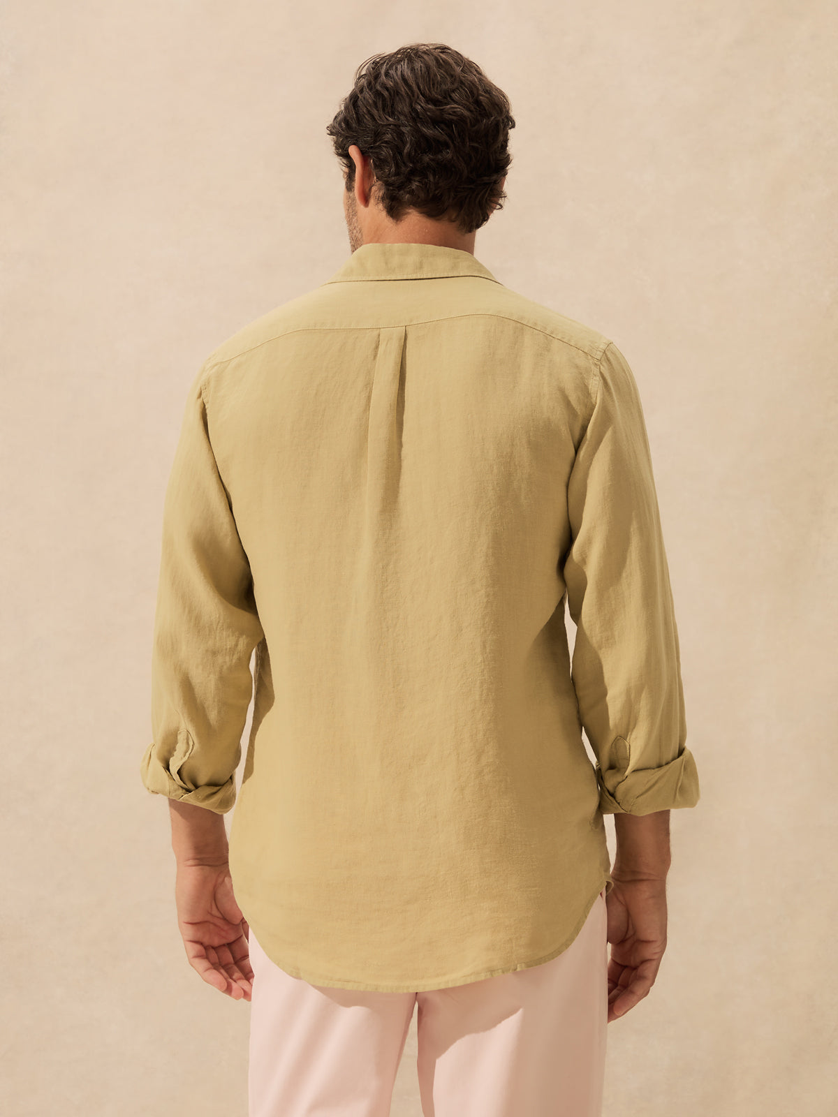 A man viewed from behind wearing a khaki linen long-sleeve button-up shirt standing against a textured beige wall.