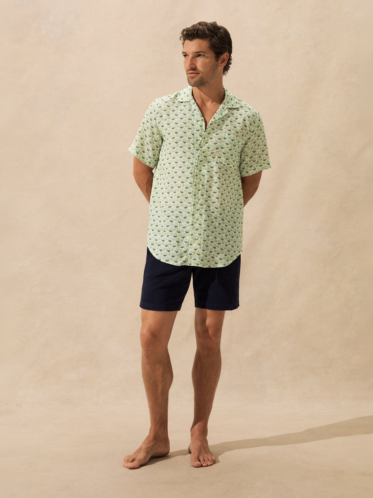 A man stands barefoot against a warm beige textured backdrop, wearing a light green short-sleeve button-up shirt with a small repeating dark pattern and dark navy shorts.