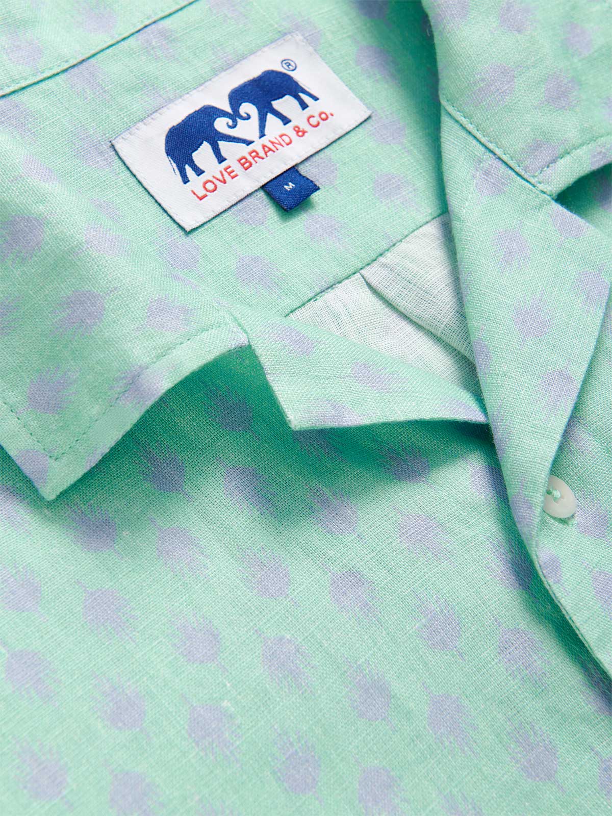Detailed view of Men's Palm Days Arawak Linen Shirt. Pastel green short-sleeved linen shirt with front pocket and subtle purple palm pattern.