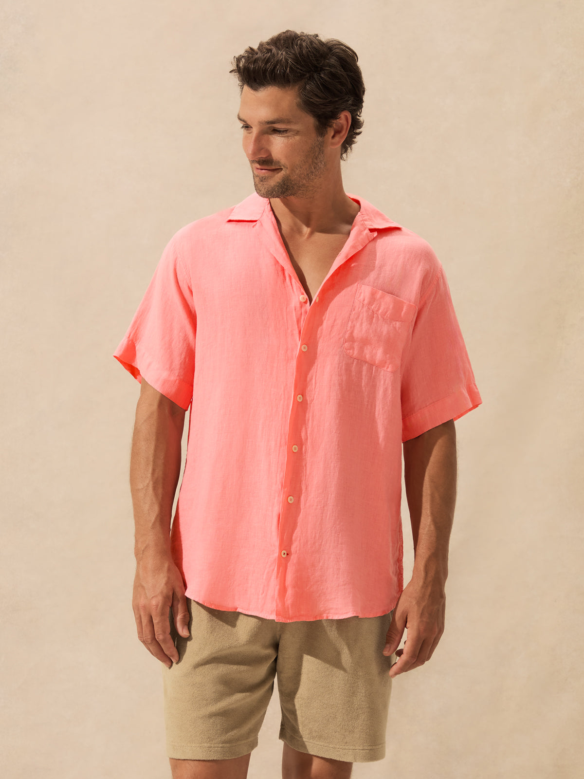 A man wearing a coral pink short-sleeve button-up shirt and beige shorts, standing against a plain beige background.