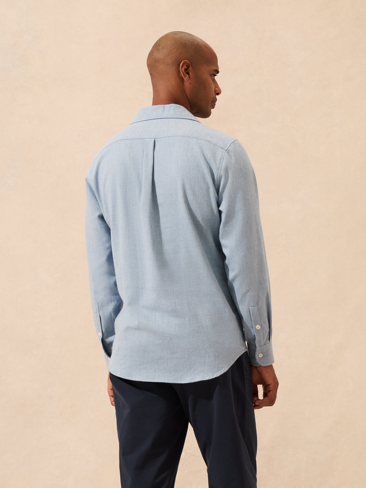 A man shown from the back wearing a light blue long-sleeve button-up shirt and dark navy trousers, standing against a plain beige textured background.