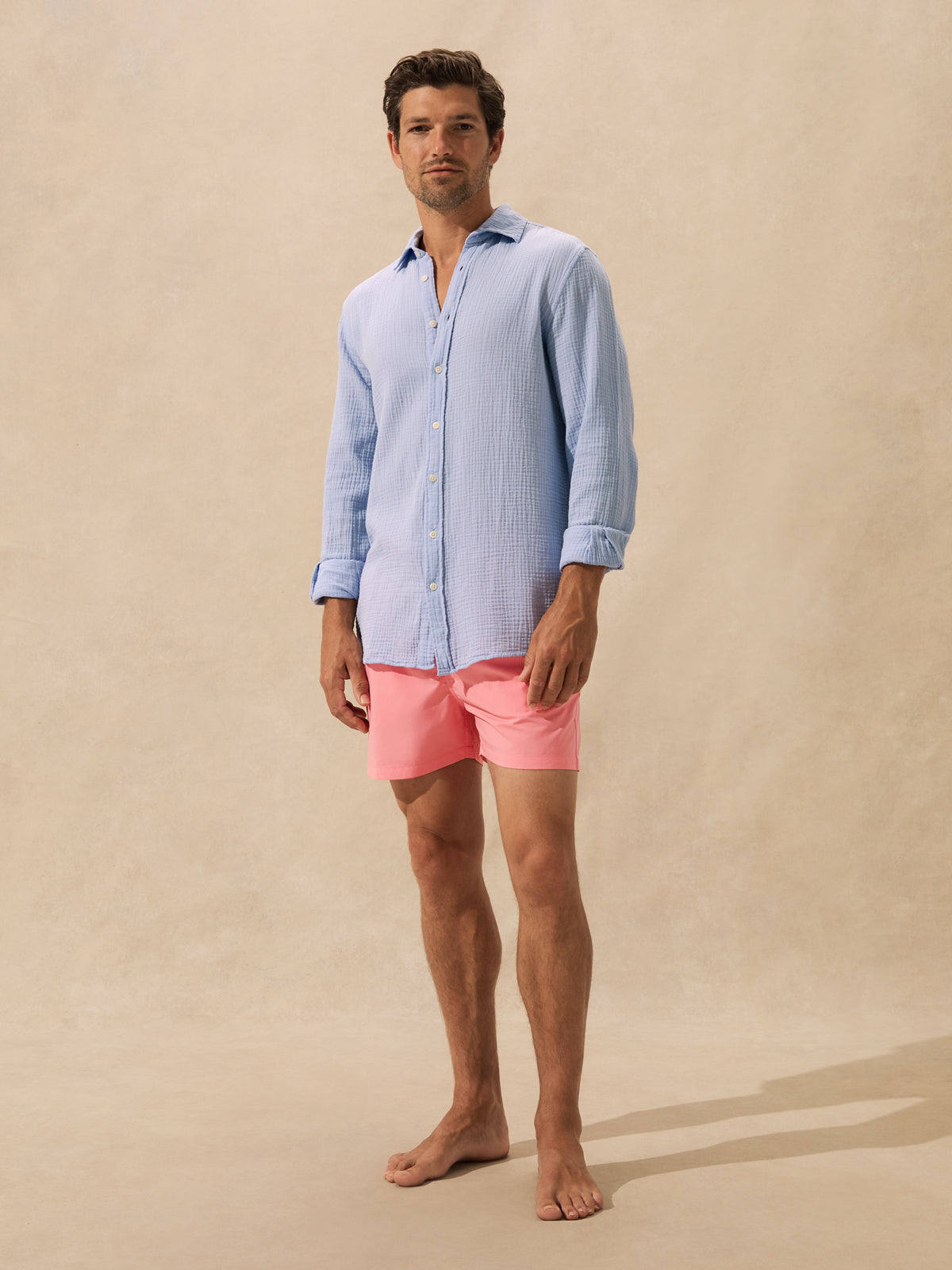 A man stands barefoot against a warm beige studio backdrop, wearing a light blue long-sleeve button-up shirt and bright pink swim trunks.