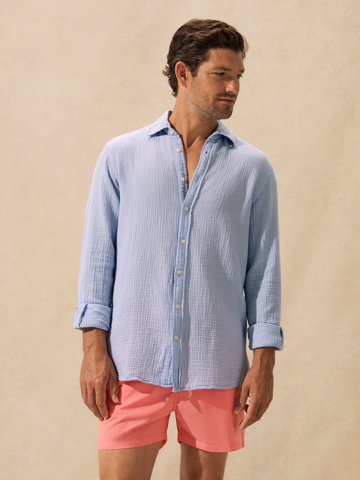 A man wearing a light blue textured long-sleeve button-up shirt and coral pink shorts, standing against a warm beige background.