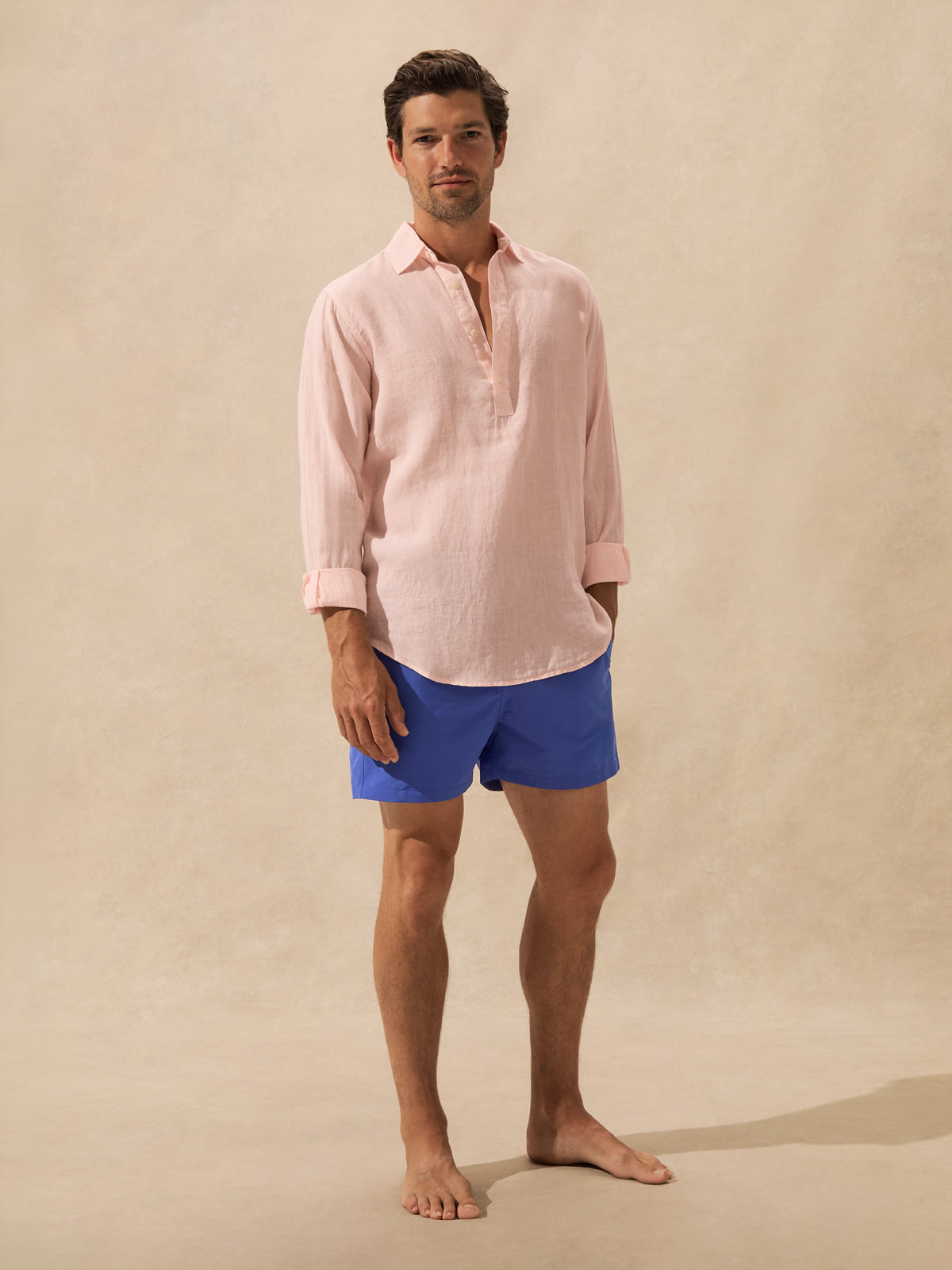 A man stands barefoot against a warm beige textured backdrop, wearing a light pink long-sleeve henley shirt and bright blue swim shorts.