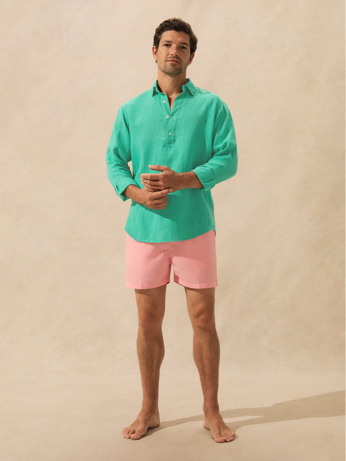 A man stands barefoot against a warm beige textured backdrop, wearing a mint green long-sleeve button-up shirt and light pink swim shorts.