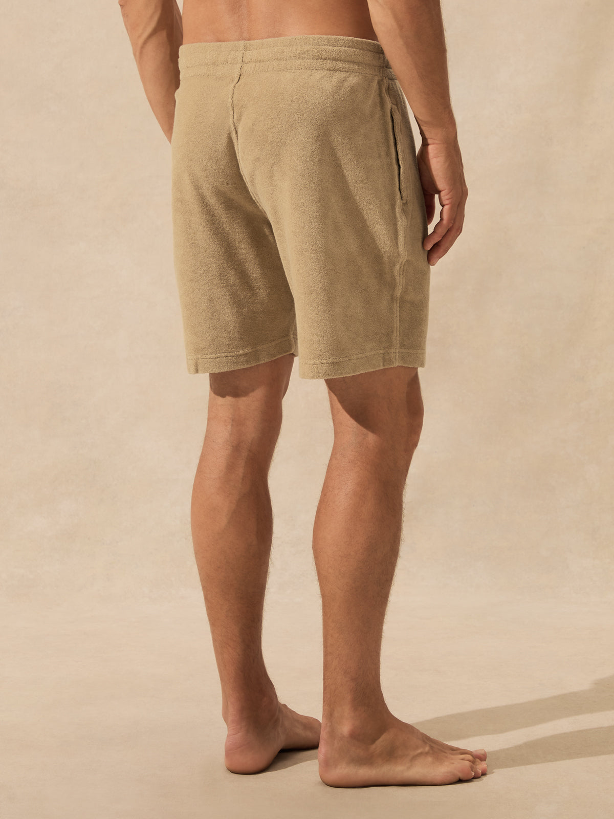 A man wearing beige drawstring shorts, shown from the back against a textured beige studio backdrop.