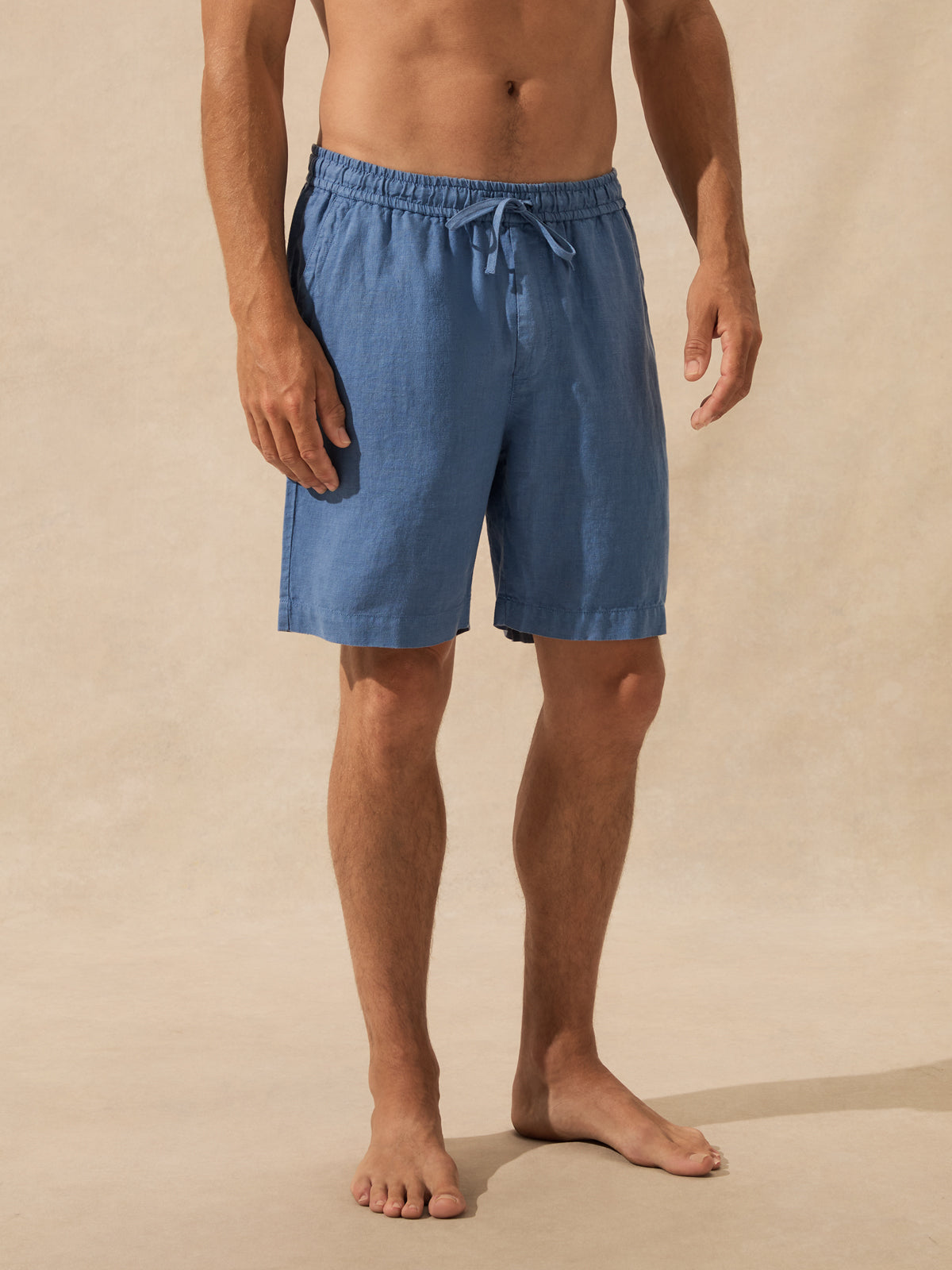 A man wearing blue drawstring shorts, shown from the waist down against a warm beige textured studio backdrop.