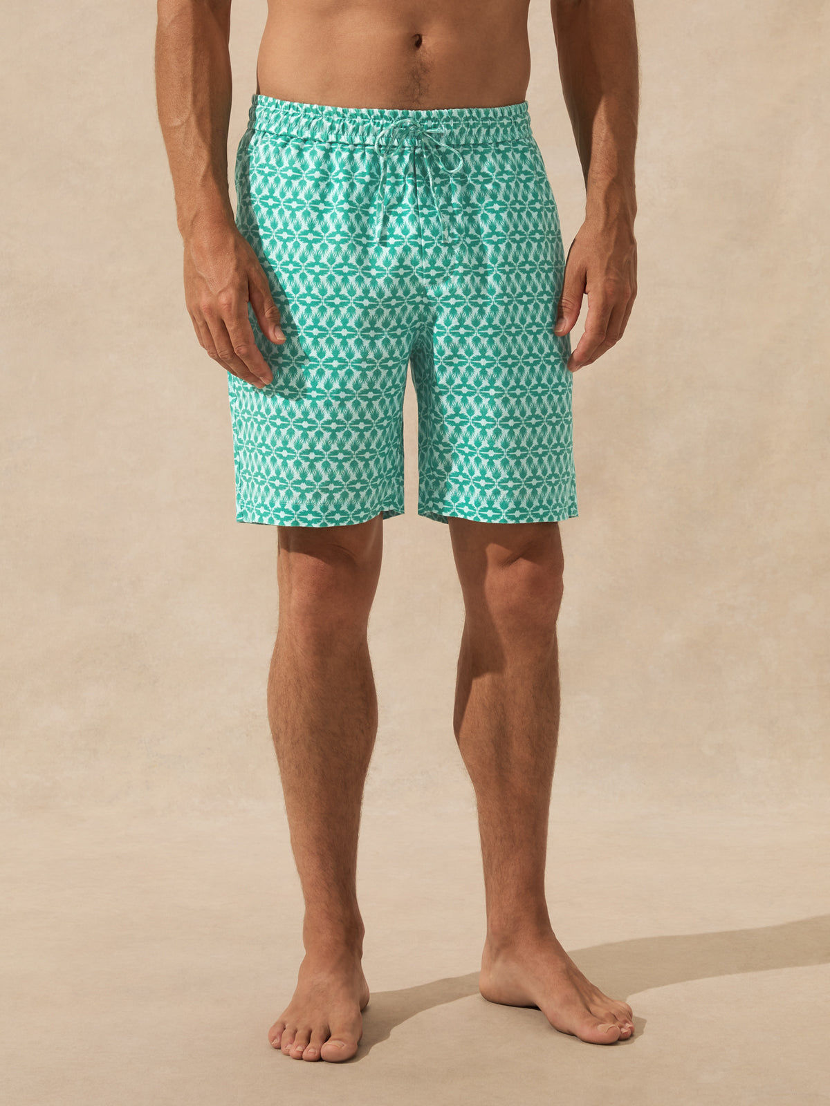 A man wearing teal and white patterned swim trunks, standing barefoot against a warm beige textured background.