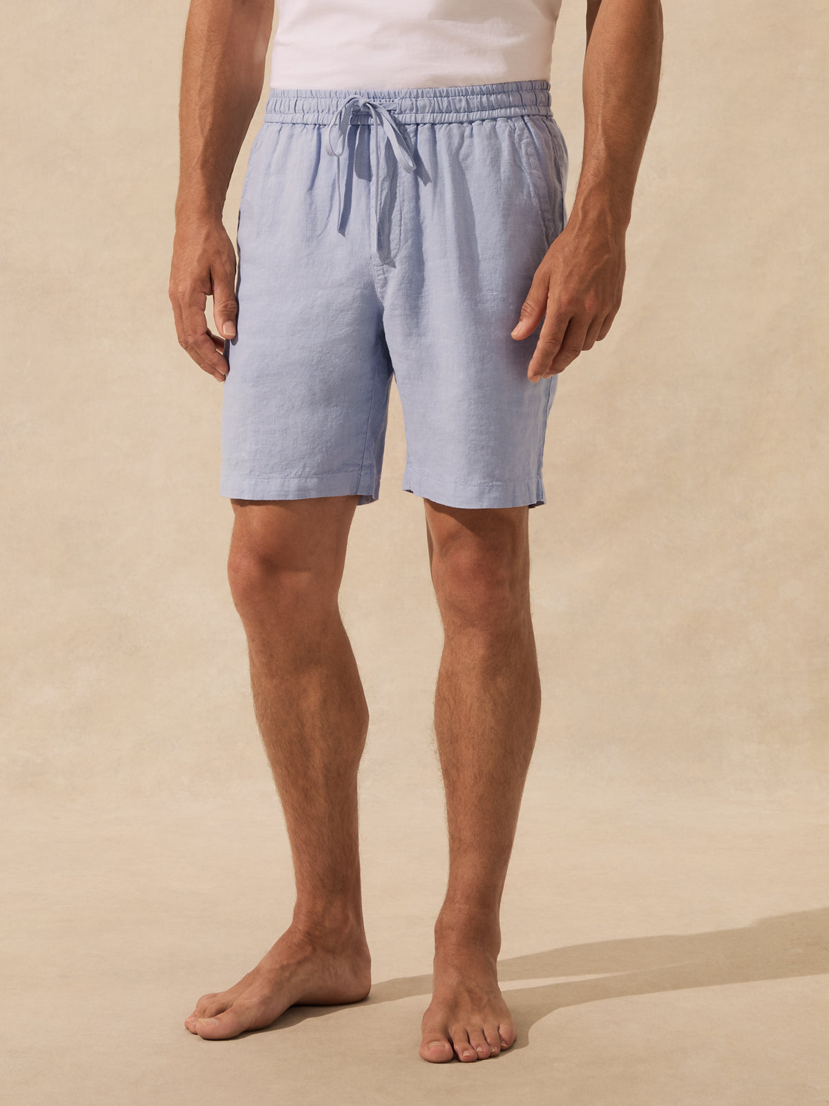 A man wearing light blue drawstring shorts standing barefoot against a warm beige textured studio background.