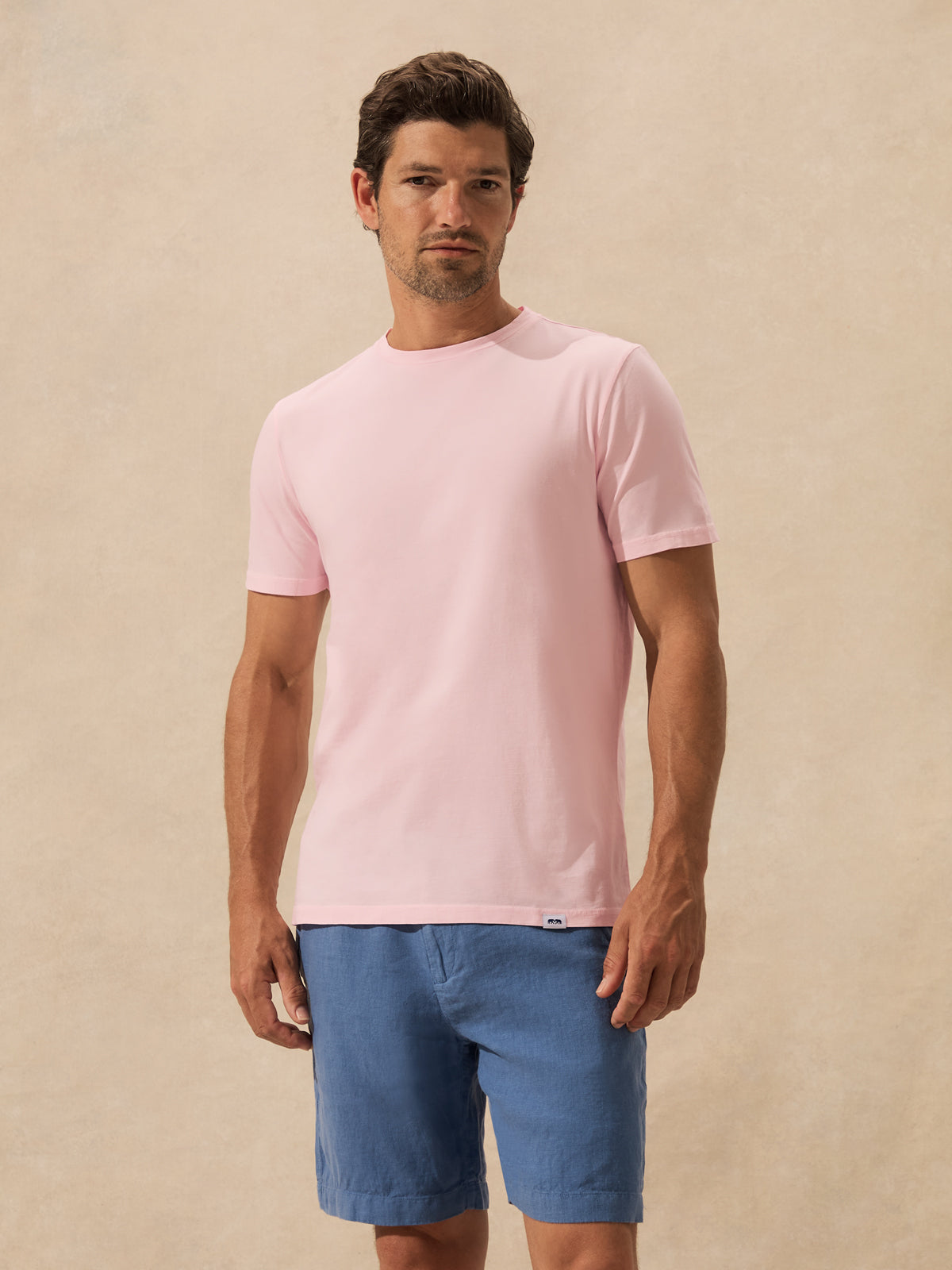 A man wearing a light pink crew-neck t-shirt and blue shorts, standing against a textured beige background.