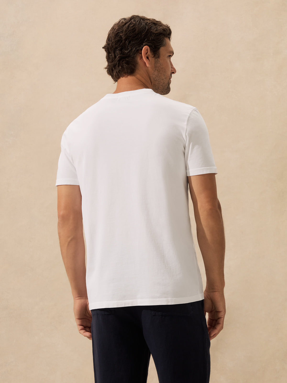 A man shown from the back wearing a plain white short-sleeve crew-neck t-shirt and dark navy trousers, standing against a textured beige studio background.