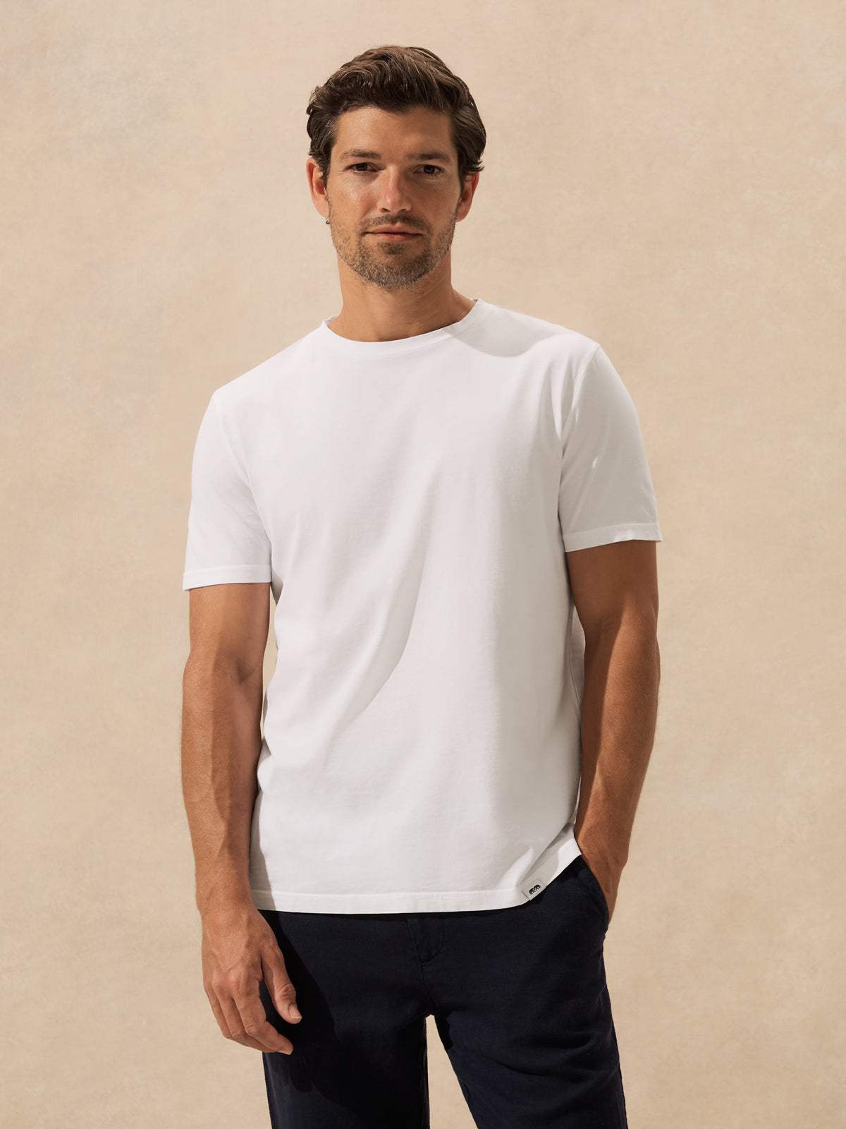A man wearing a plain white short-sleeve crewneck t-shirt and dark navy pants, standing against a textured beige background.