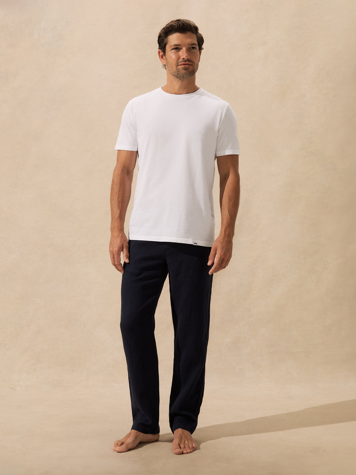 A man stands barefoot against a warm beige textured studio backdrop, wearing a plain white short-sleeve crew-neck T-shirt and dark navy pants.