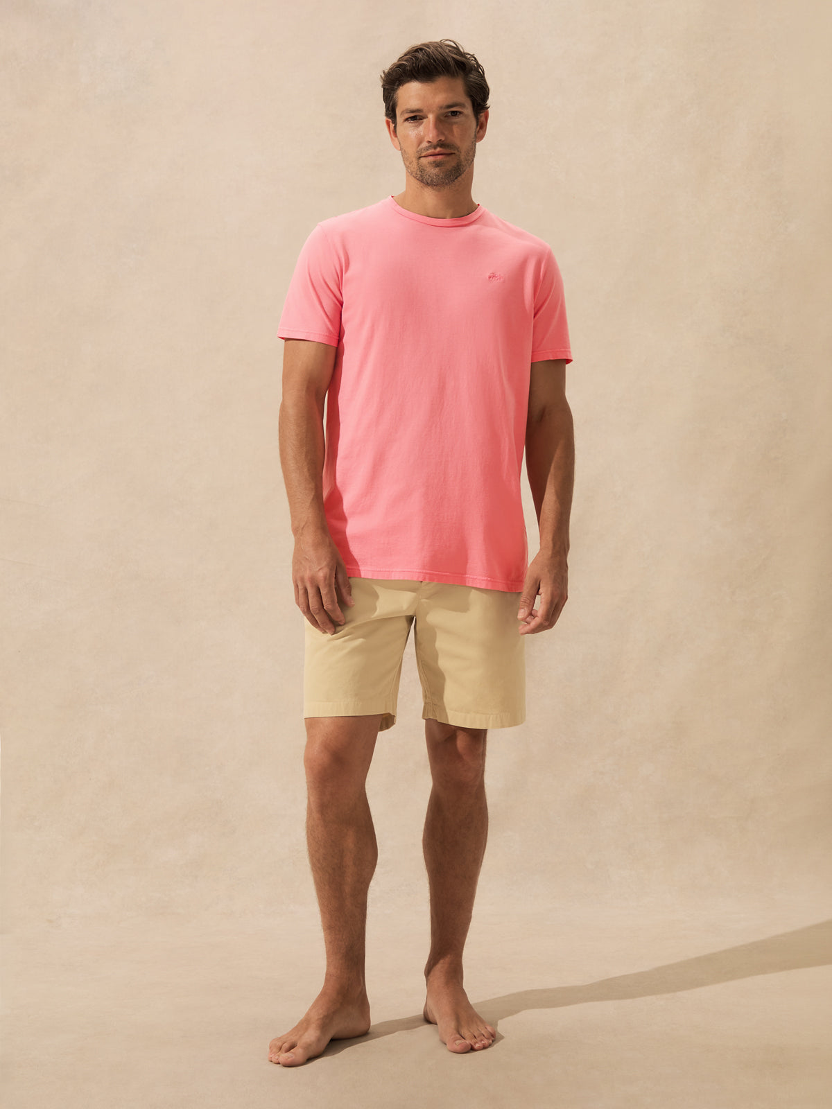 A man wearing a coral pink crewneck t-shirt and beige shorts, standing barefoot against a textured beige studio backdrop.