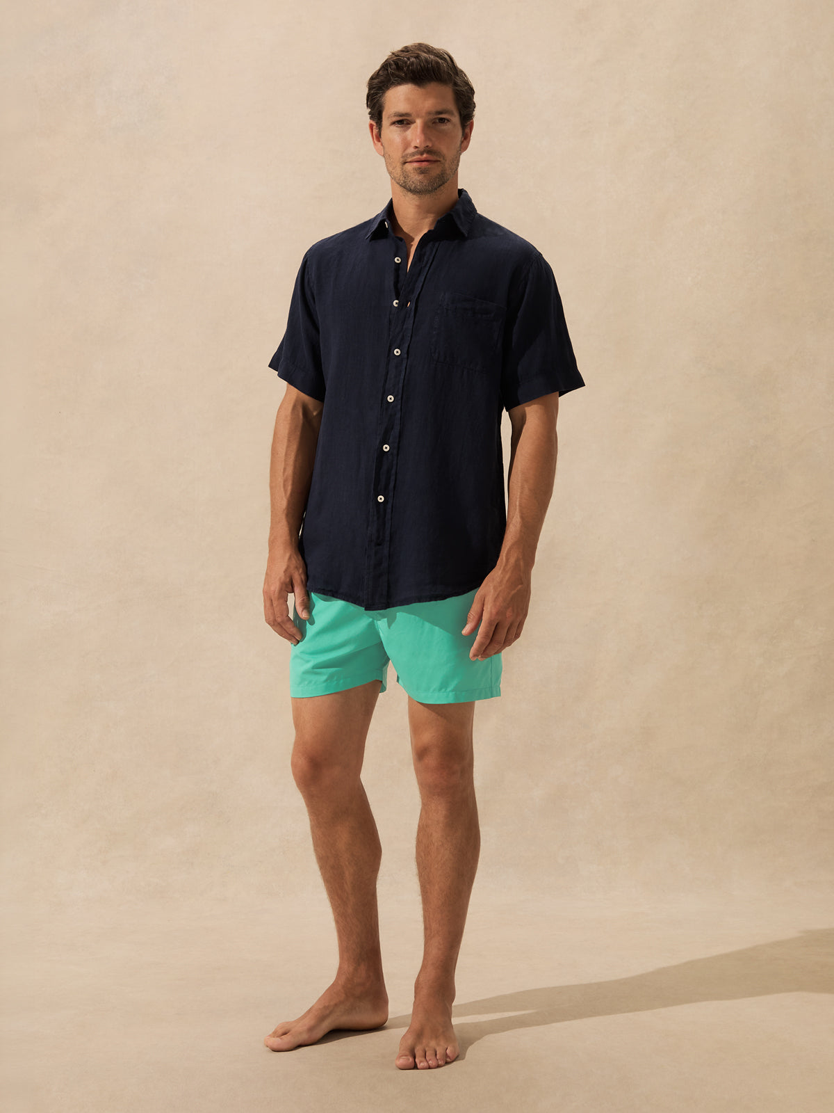 A man stands barefoot against a warm beige textured studio backdrop, wearing a dark navy short-sleeve button-up shirt and bright aqua-green shorts.