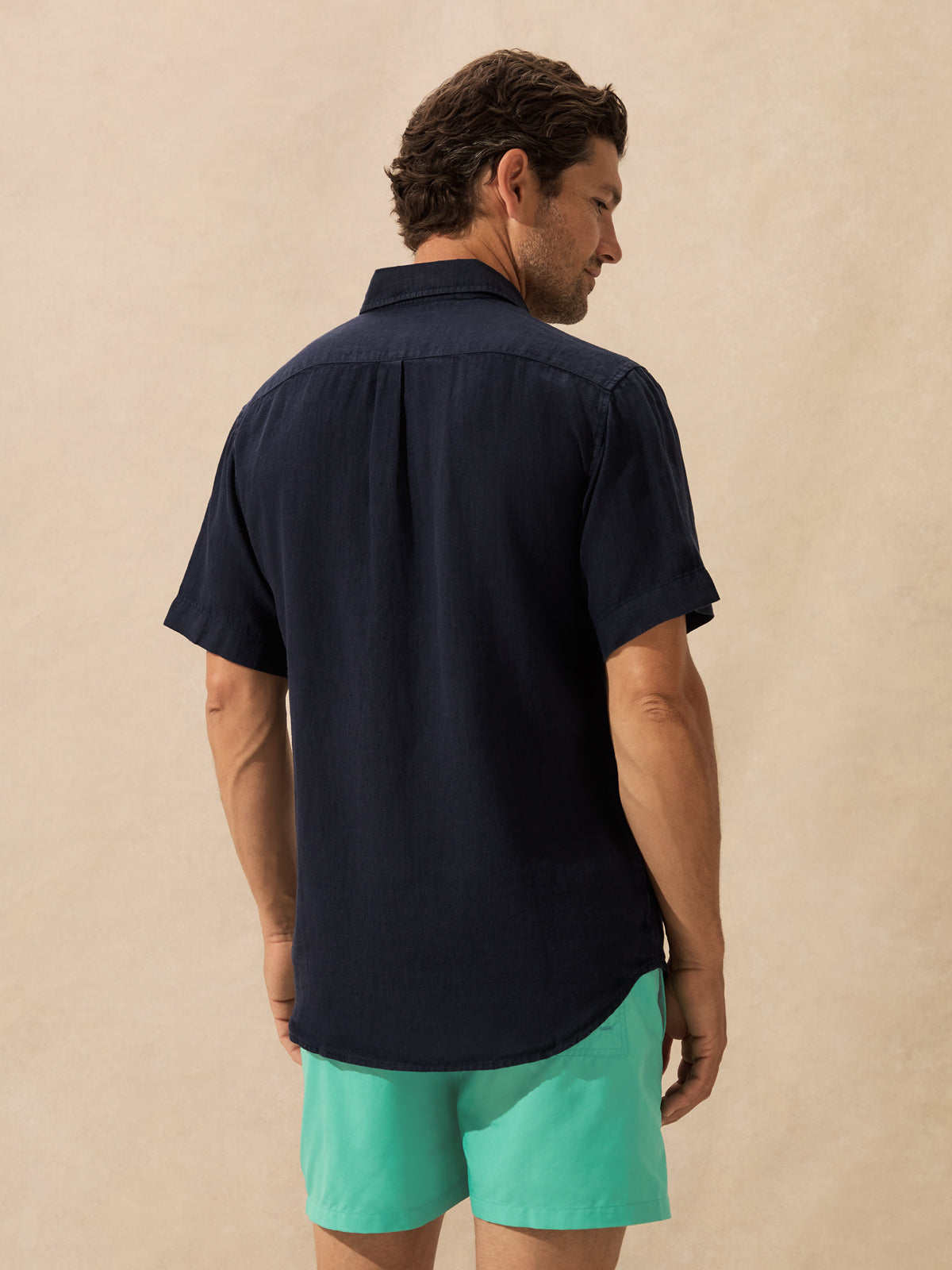 A man shown from the back wearing a dark navy short-sleeve button-up shirt and bright mint green shorts, standing against a textured beige background.