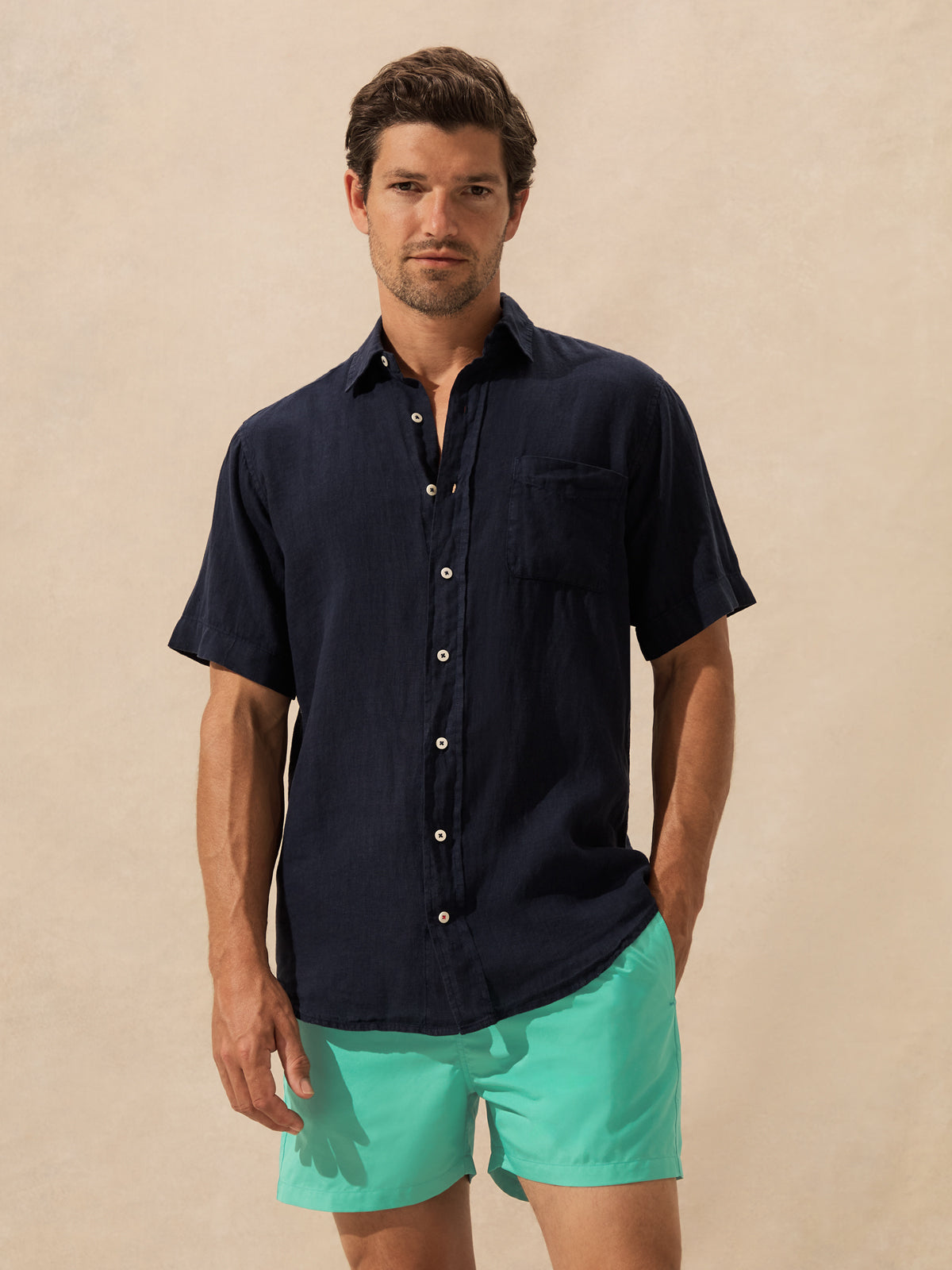 A man wearing a dark navy blue short-sleeve button-down shirt and bright turquoise shorts, standing against a textured beige background.