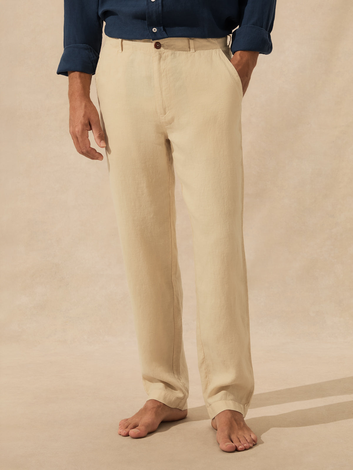 A person wearing straight-leg beige trousers, standing barefoot against a warm beige textured studio backdrop.
