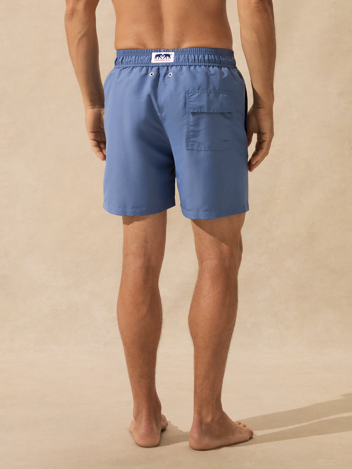 A man shown from the back wearing light blue drawstring swim trunks with a side pocket, standing barefoot against a warm beige textured studio background.