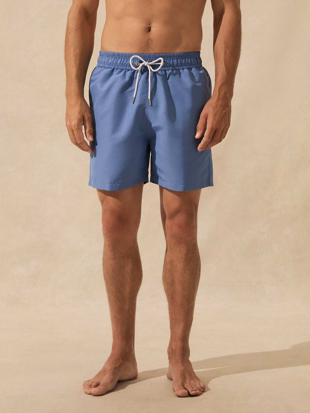 A man wearing blue swim trunks with a white drawstring, standing barefoot against a warm beige textured background.