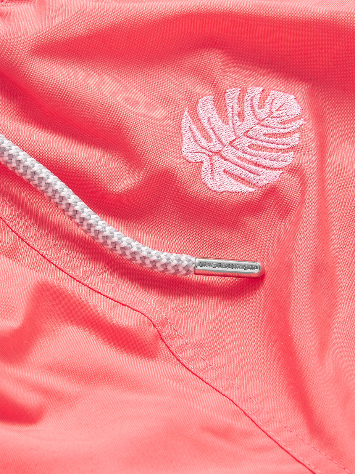 Close-up view of Boys Deliciosa Embroidered Staniel Swim Shorts featuring a Coral Rose base with pink monstera deliciosa leaf embroidery and a white drawstring.