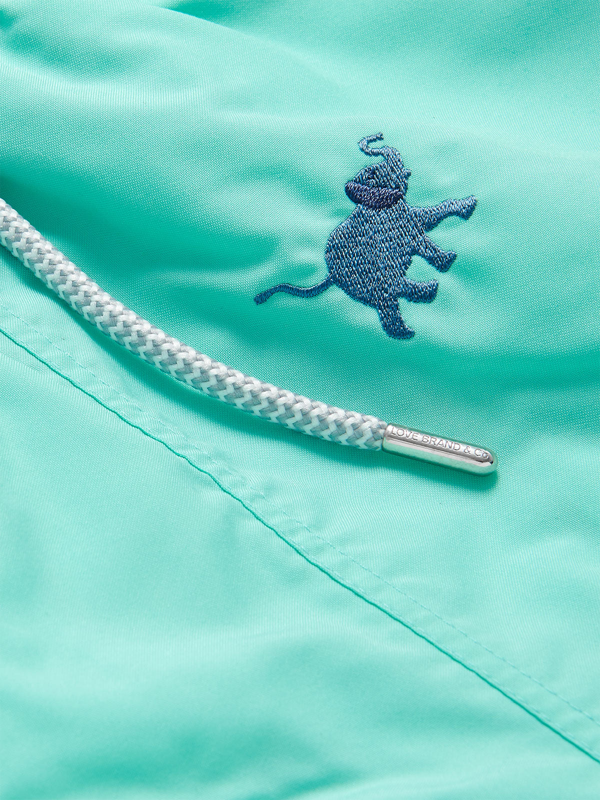 Close-up of Men's Elephants Galore Embroidered Staniel Swim Shorts in Cay Green featuring a Deep Blue embroidered elephant and a braided drawstring with a branded metal tip.